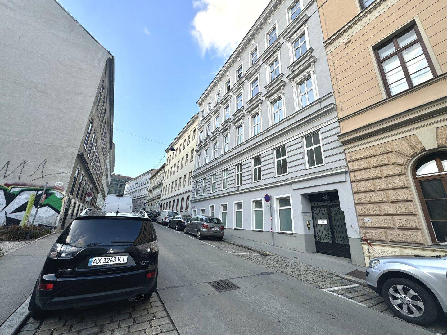 Lange Häuserzeile in einer städtischen Straße mit geparkten Autos und einem Gebäude mit Graffiti.