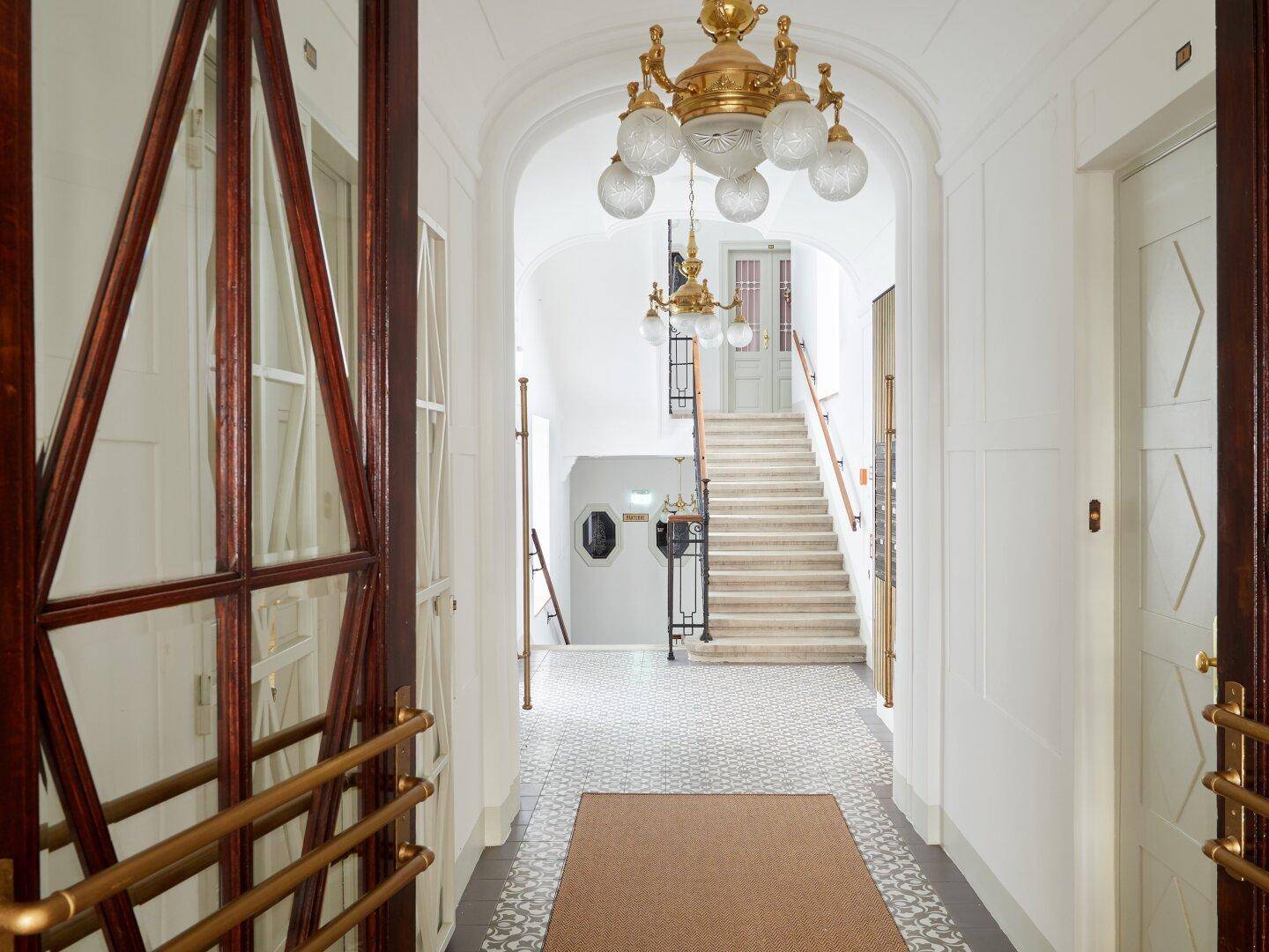 Heller Eingangsbereich mit hohen Decken, eleganten Kronleuchtern und Zugang zur Treppe.