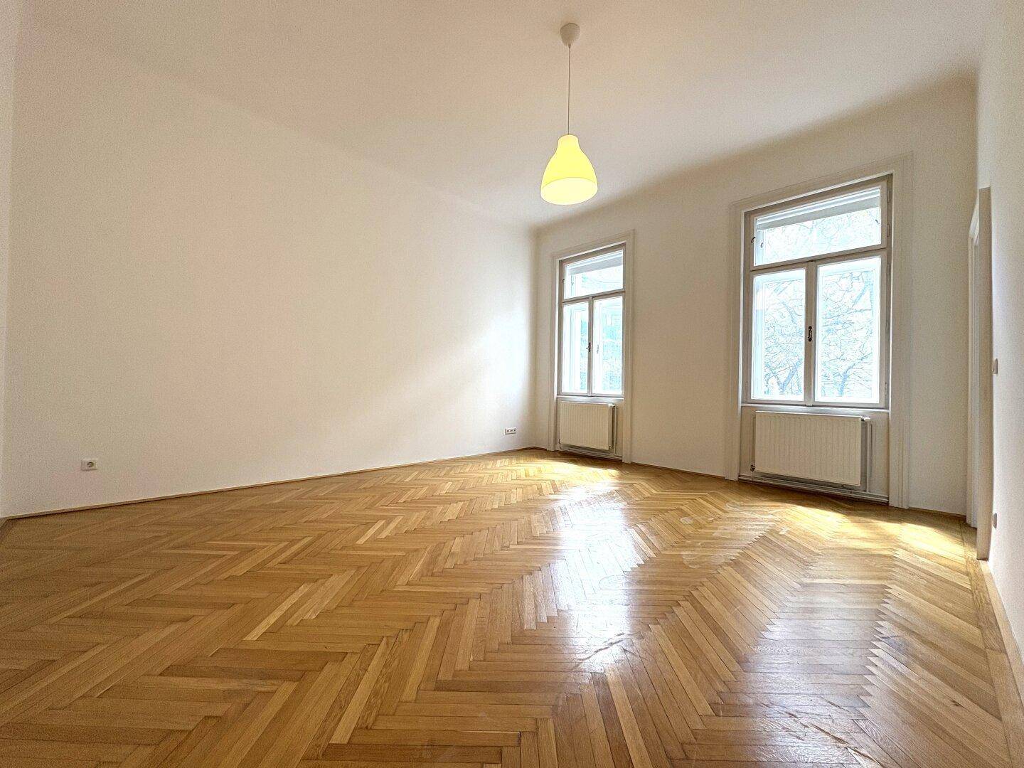 Geräumiges Wohnzimmer mit viel Tageslicht, klassischem Fischgrätparkett und schlichten weißen Wänden.