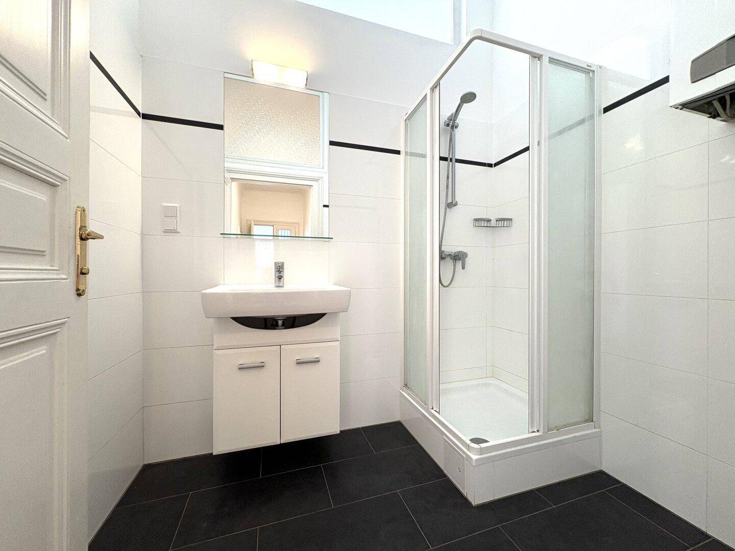 Modernes Badezimmer mit Duschkabine, weißem Waschbecken und dunklen Bodenfliesen.
