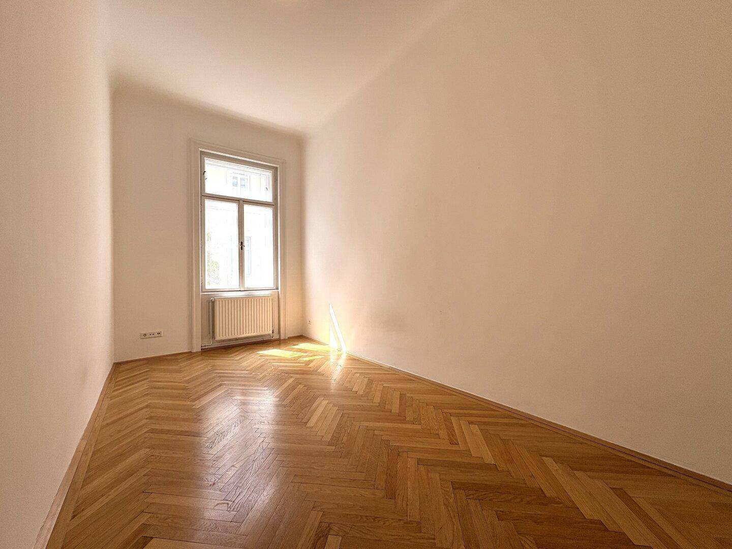 Helles Zimmer mit einem Fenster, Fischgrätparkettboden und weiß gestrichenen Wänden.