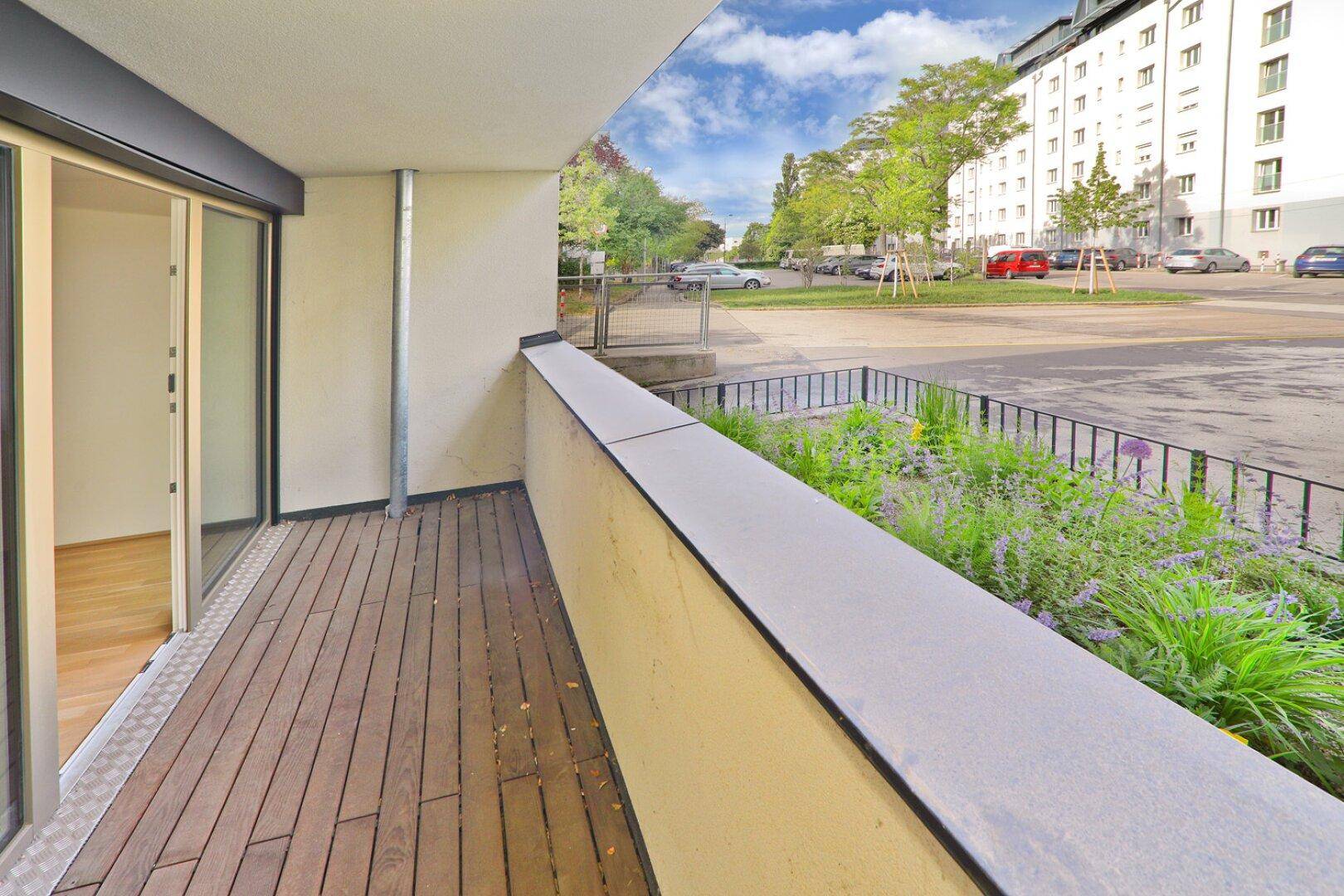 Breiter Balkon mit Holzboden und Blick auf die begrünte Umgebung und die Straße.
