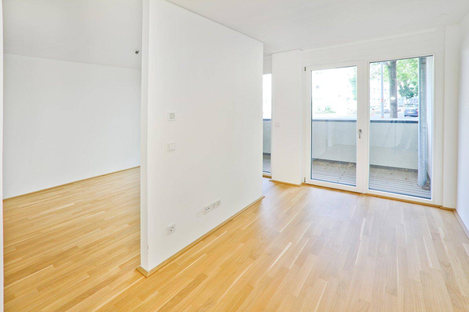 Heller Wohnraum mit Holzboden und Balkonzugang, durch eine Trennwand strukturiert.