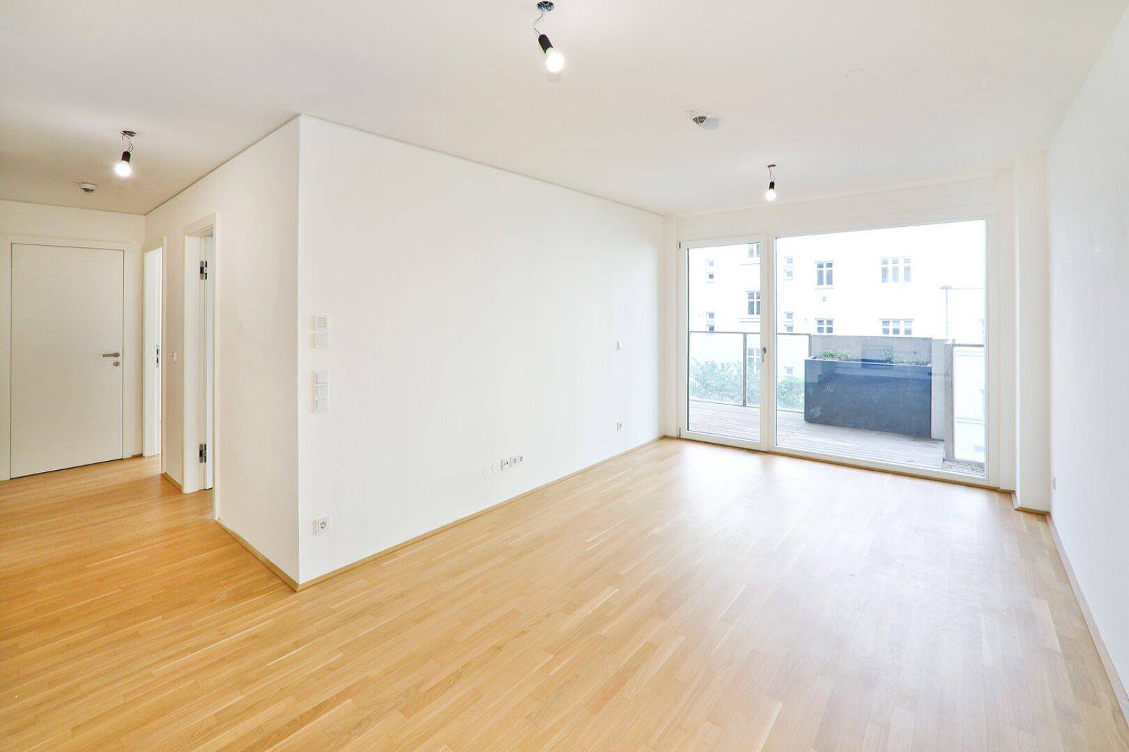 Heller, leerer Wohnraum mit Parkettboden und Zugang zu einem Balkon.
