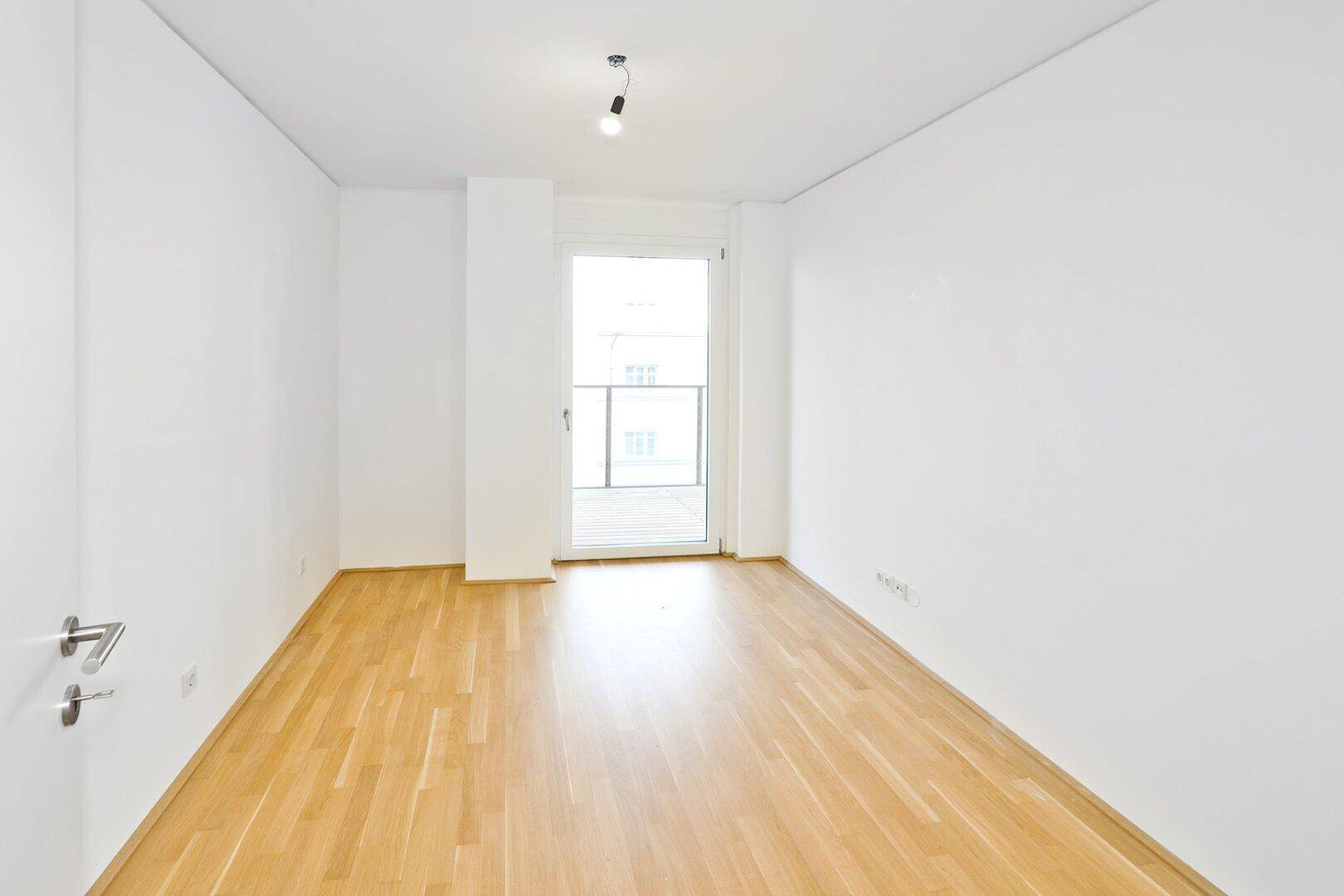 Leeres Zimmer mit Parkettboden und Zugang zu einem kleinen Balkon.