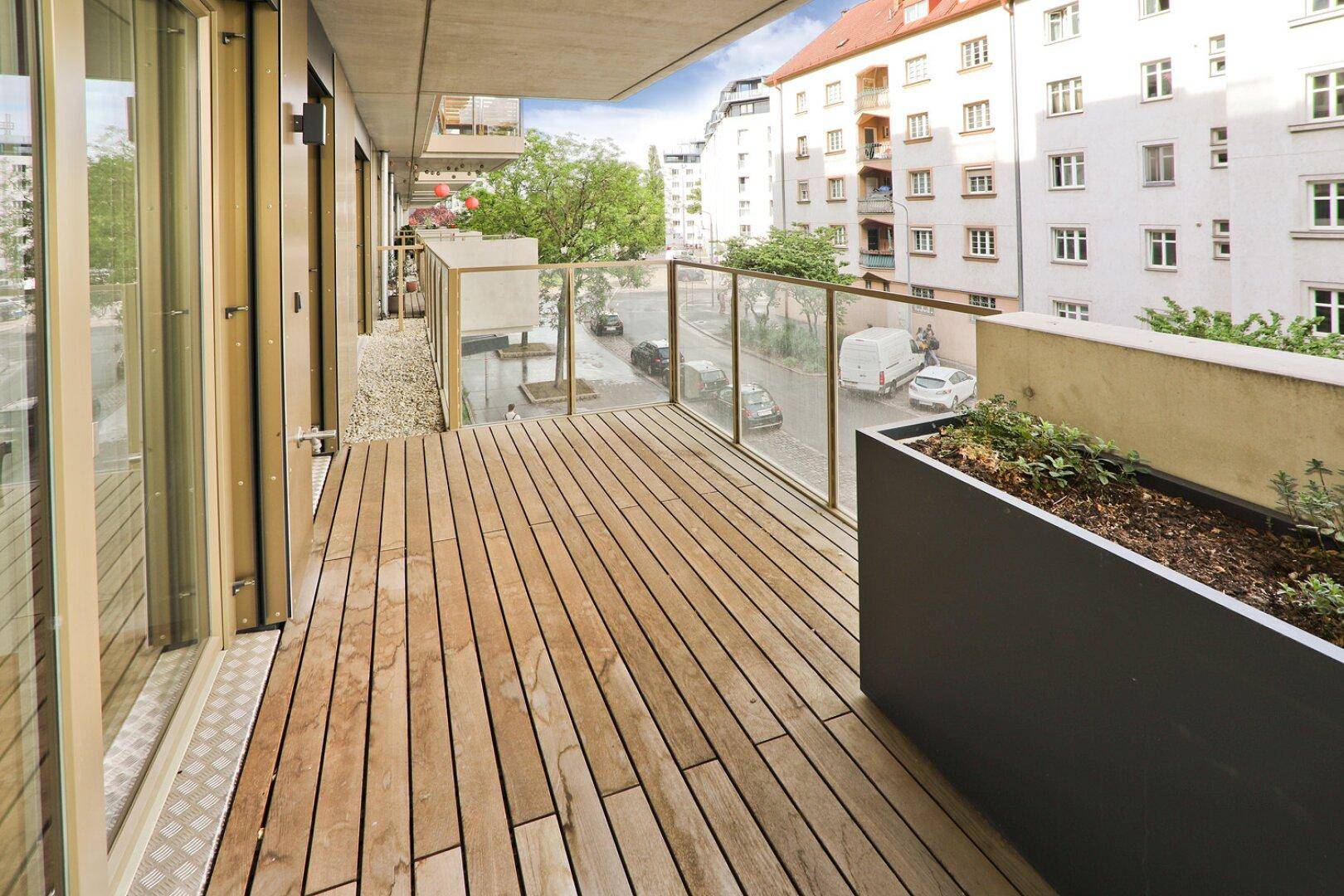 Breiter Balkon mit Holzfußboden, Glasgeländer und Blick auf die belebte Straße.
