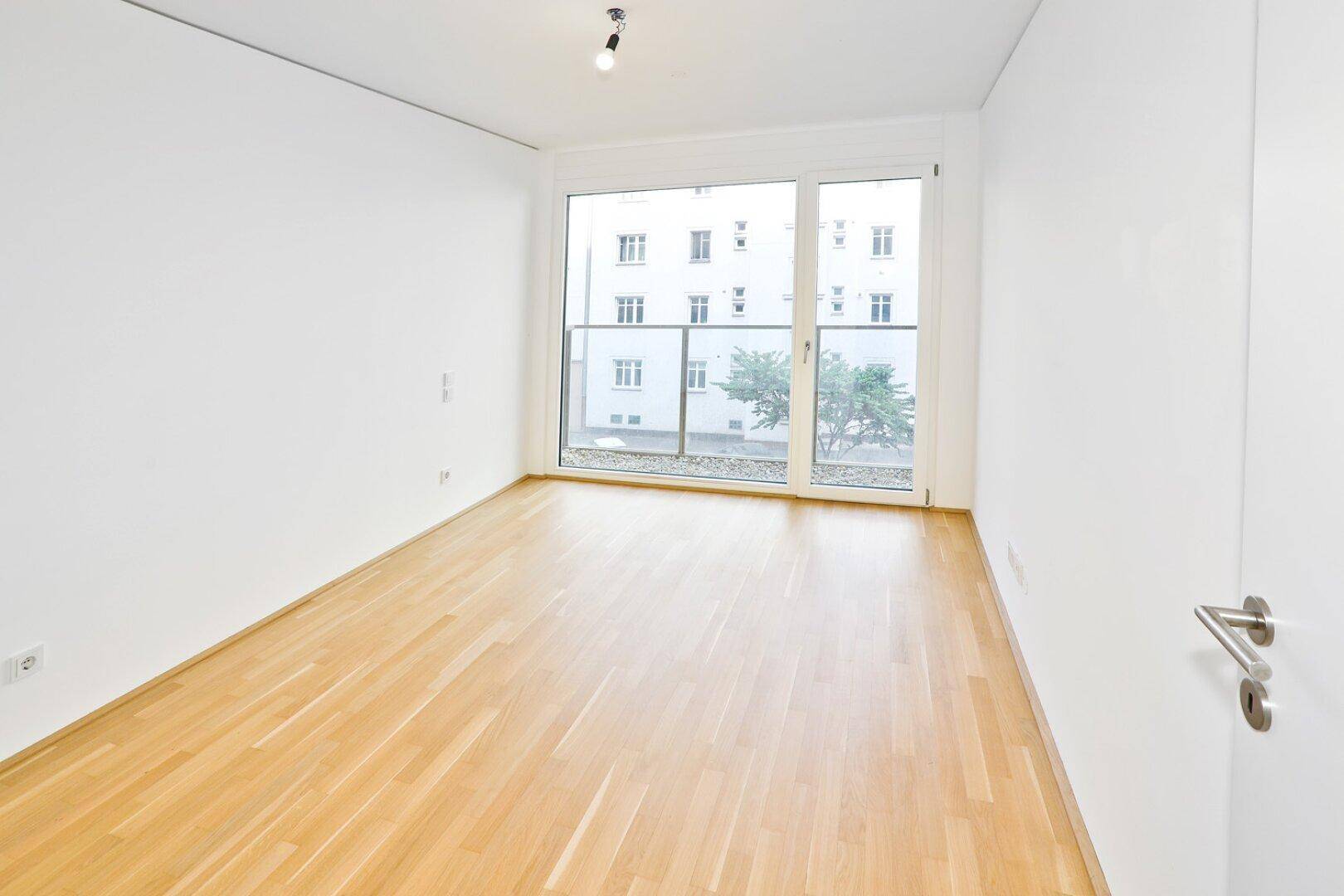 Lichtdurchflutetes Zimmer mit Parkettboden und großem Fenster, bietet einen ruhigen Ausblick.