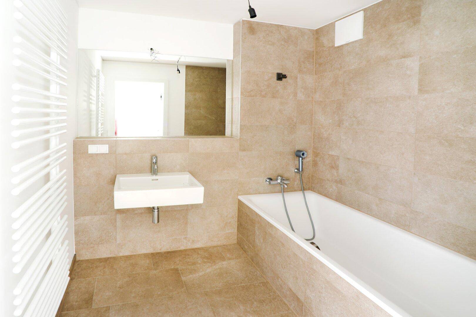 Modernes Badezimmer mit beigefarbenen Fliesen, Badewanne und beheiztem Handtuchhalter.