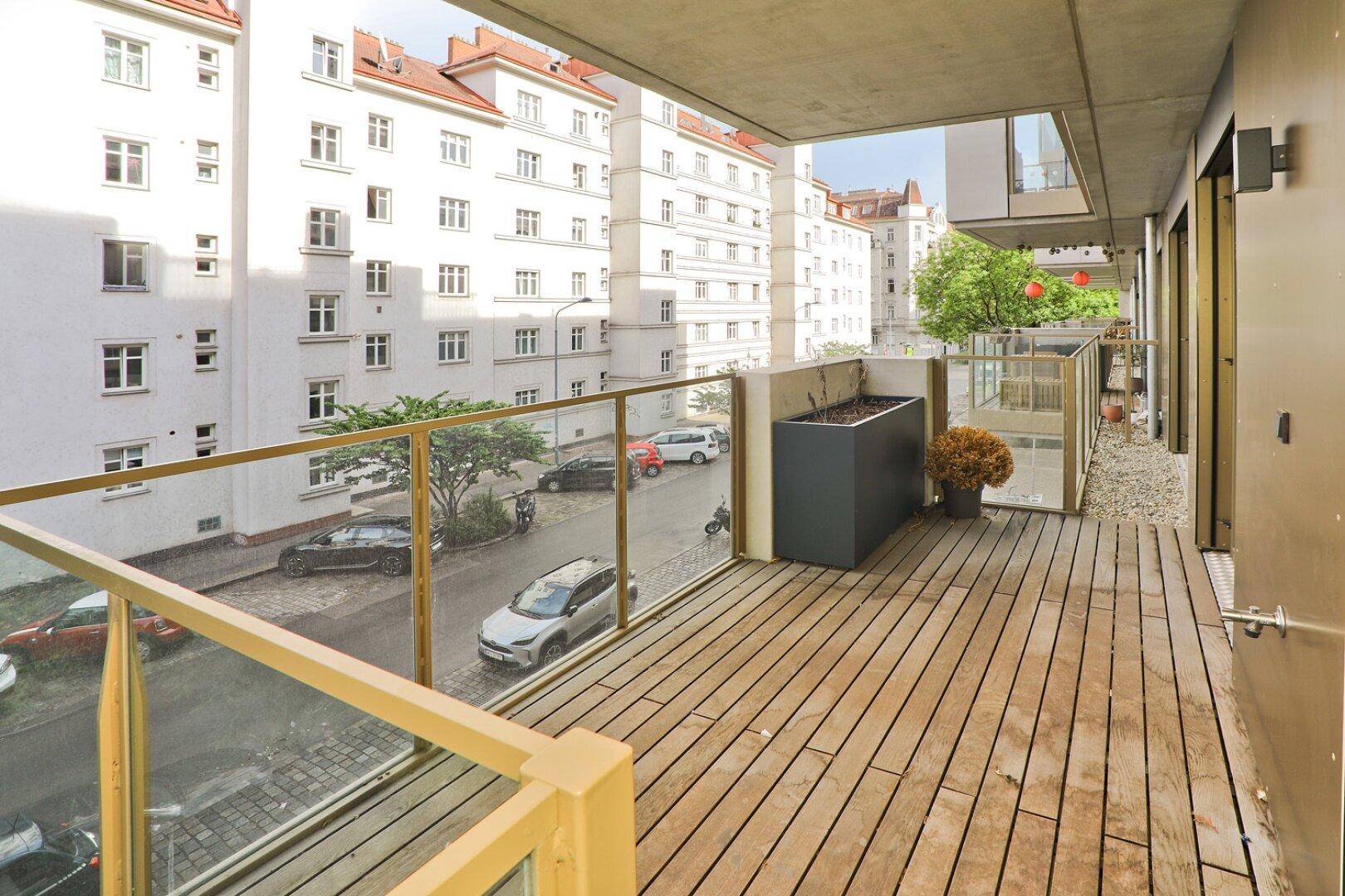 Balkon mit Holzboden und Glasgeländer, bietet Blick auf die Straße und umliegende Gebäude.