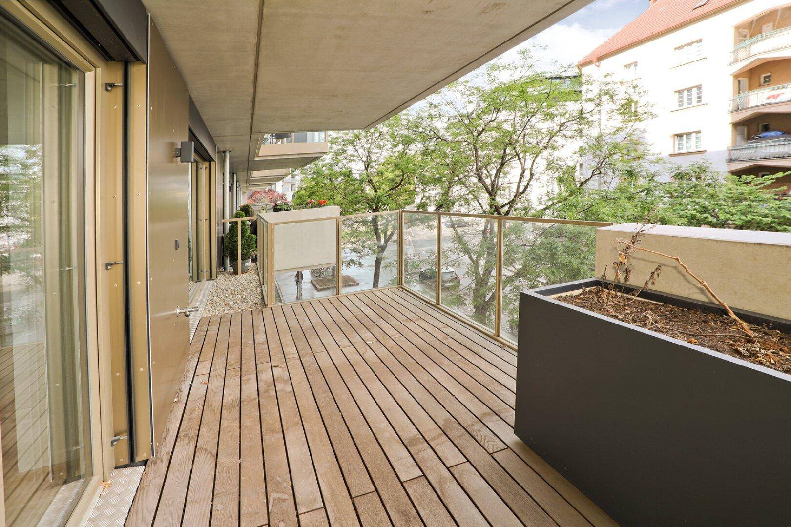 Geräumiger Balkon mit Holzdielen und Glasbrüstung, umgeben von Bäumen und Gebäuden.