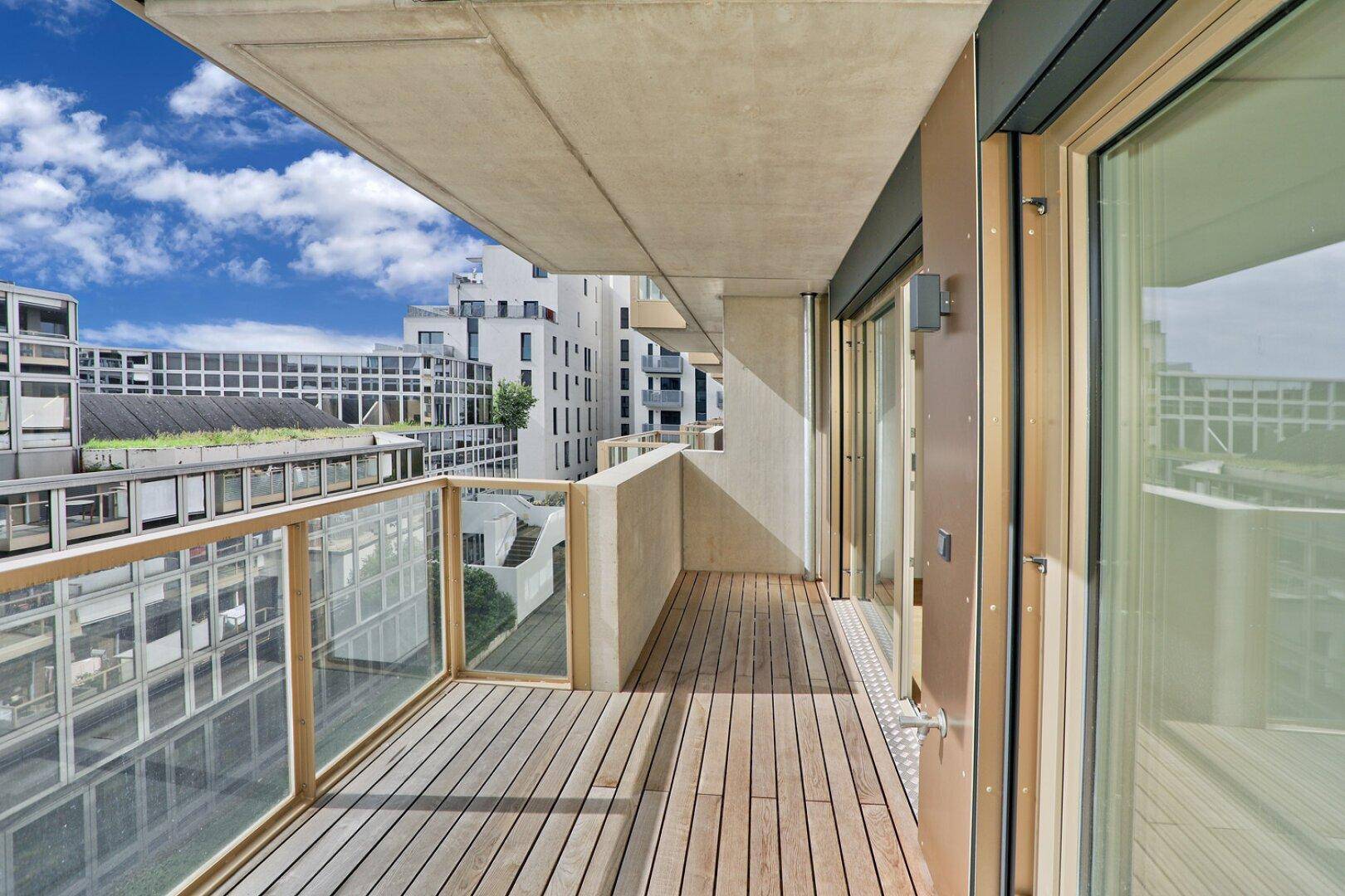 Geräumiger Balkon mit Holzbelag und Glasgeländer, bietet einen Blick auf moderne Gebäude.