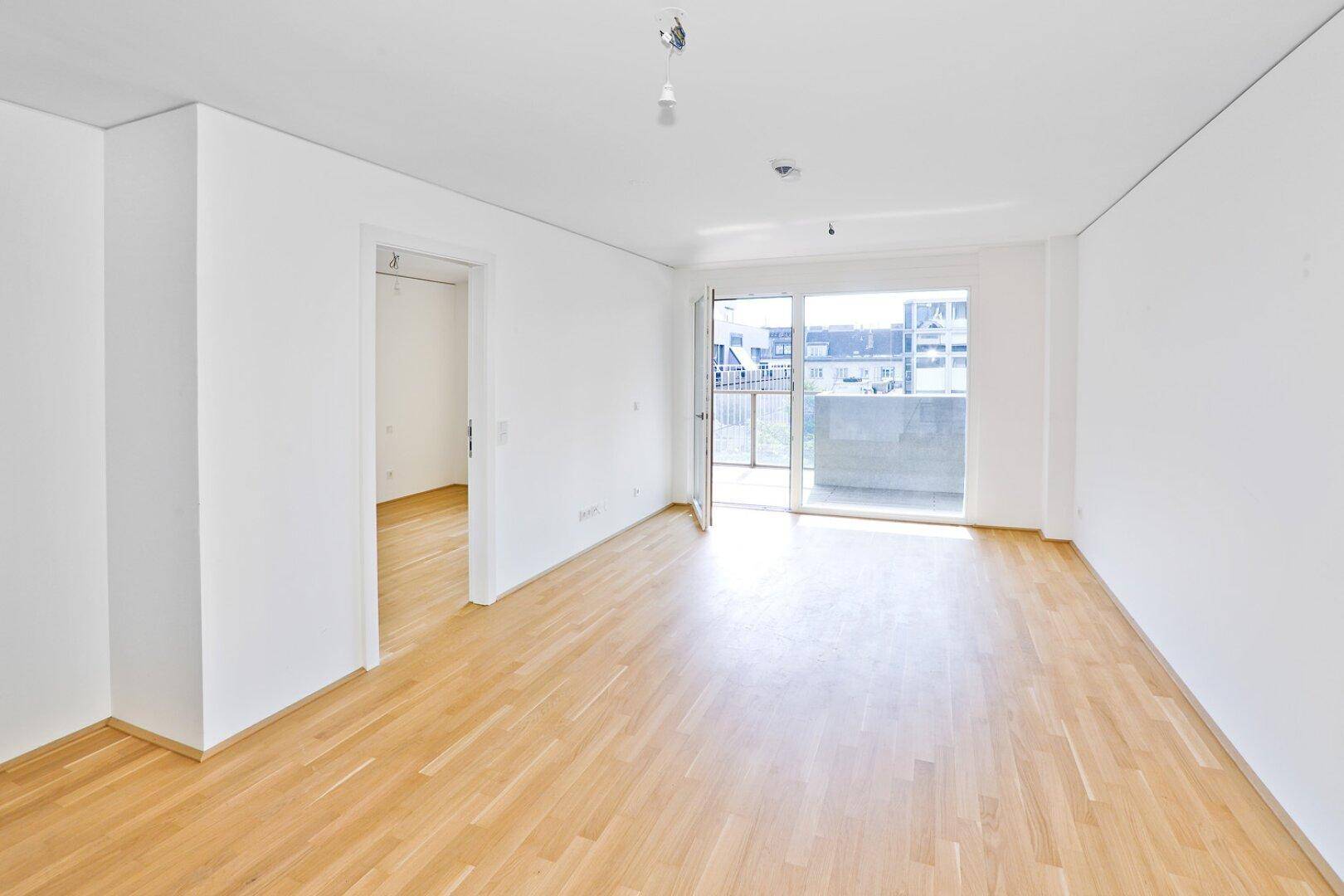 Helles, leeres Wohnzimmer mit hellem Parkettboden und Zugang zu einem Balkon.