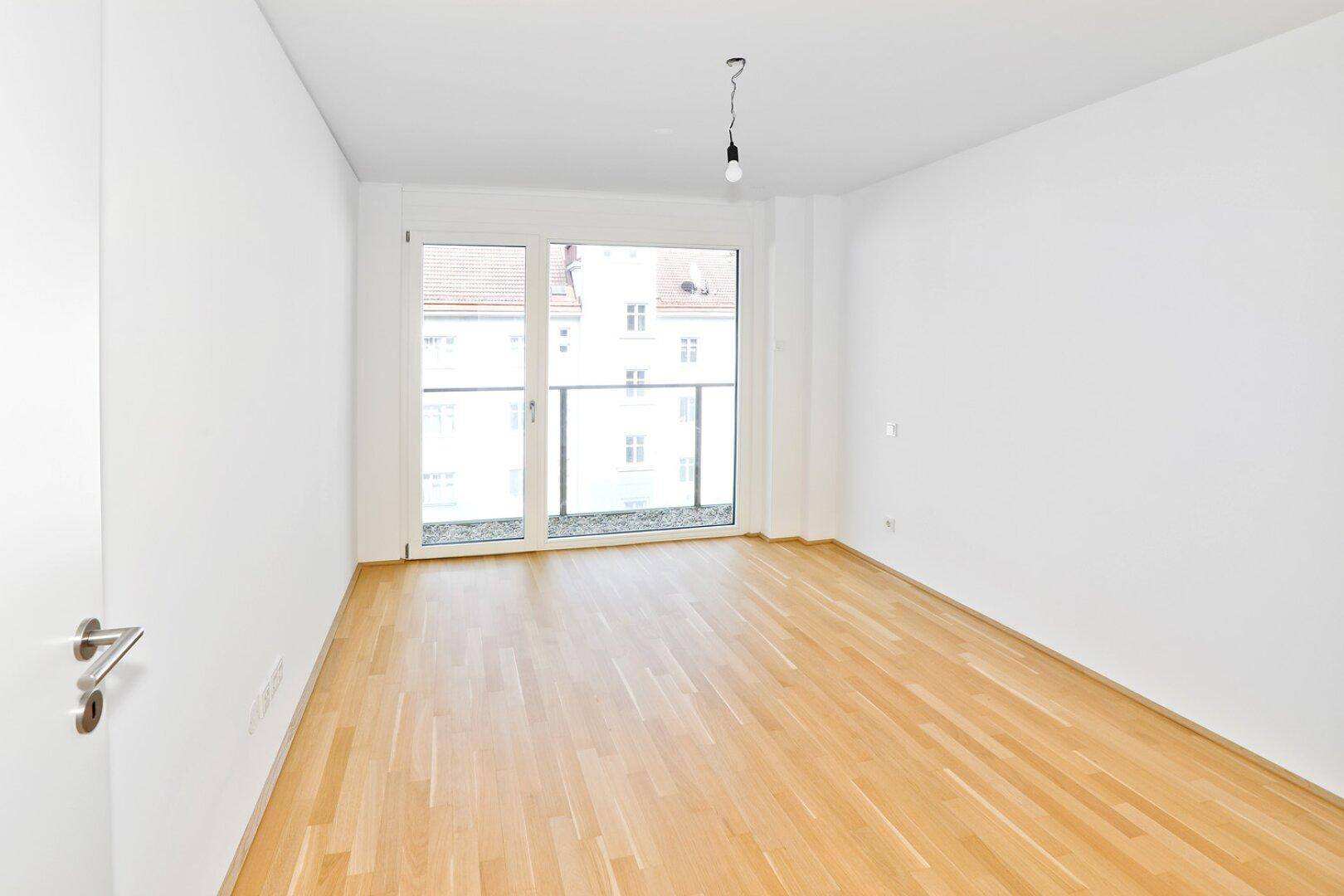 Leeres Schlafzimmer mit hellem Parkettboden und großem Fenster mit Zugang zum Balkon.