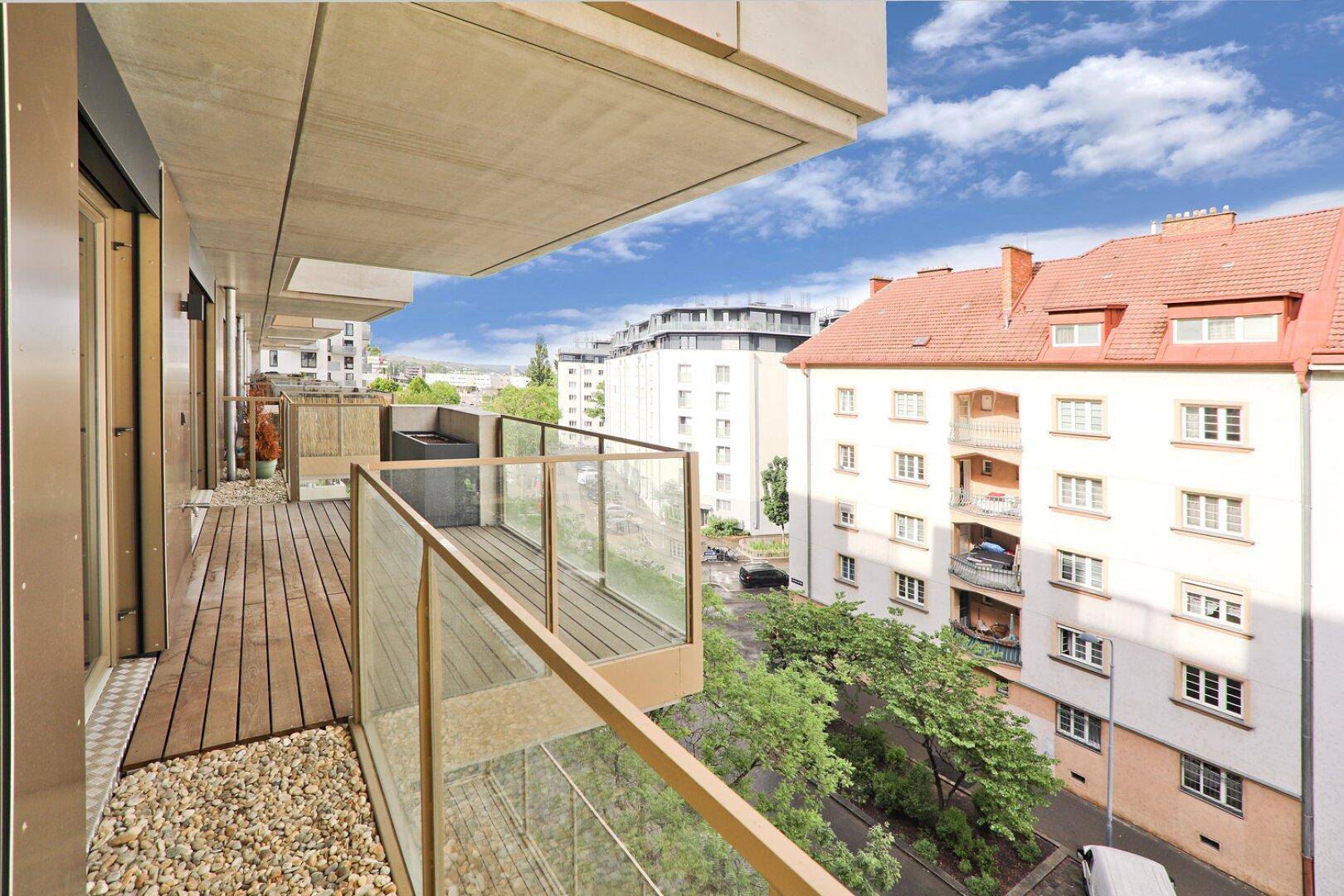 Heller Balkon mit Holzdielen und Glasbrüstung, mit Blick auf die umliegenden Gebäude und die Straße.