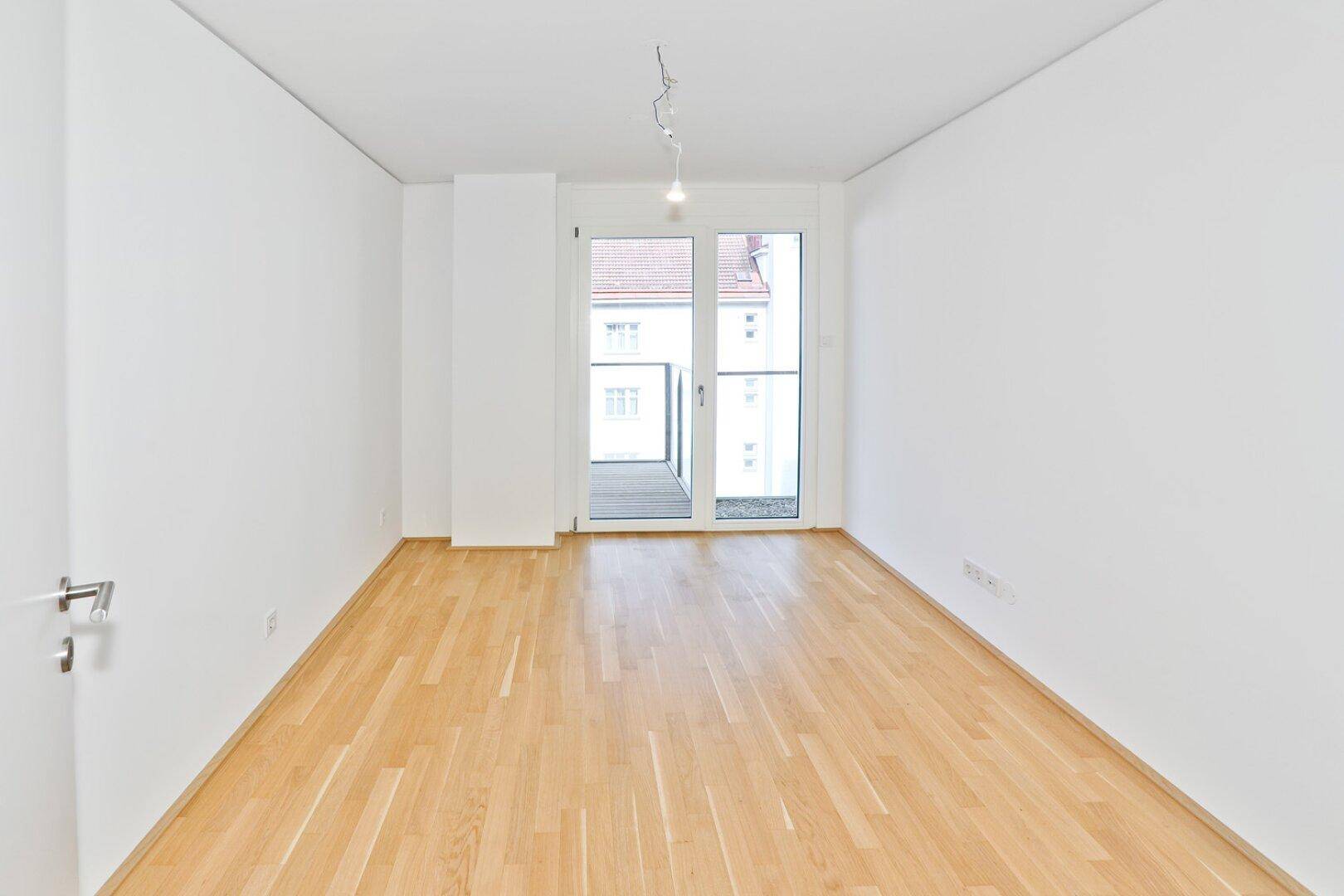 Leeres Zimmer mit hellem Parkettboden und großen Fenstern, die viel Tageslicht hereinlassen.