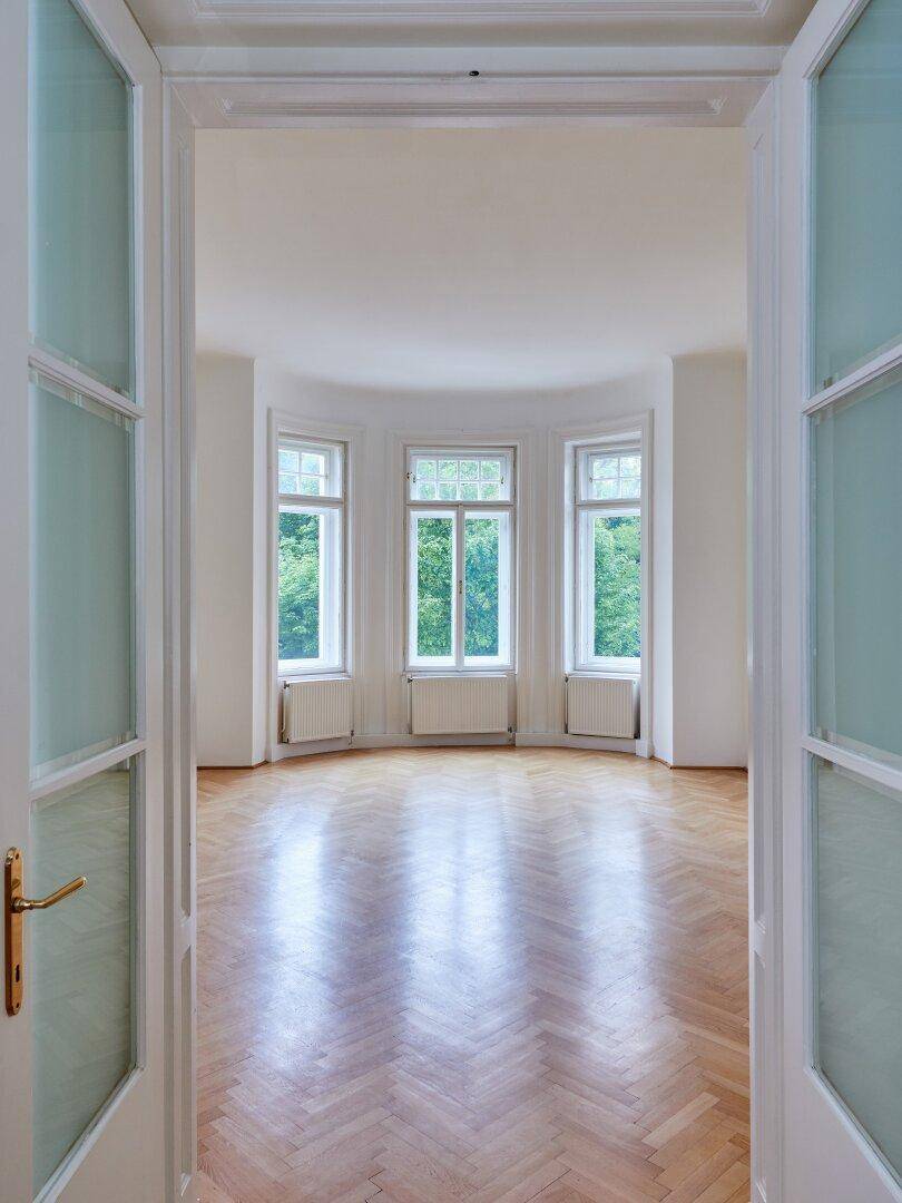 Blick durch eine offene Tür in ein helles Zimmer mit Fischgrätparkett und Erkerfenstern.