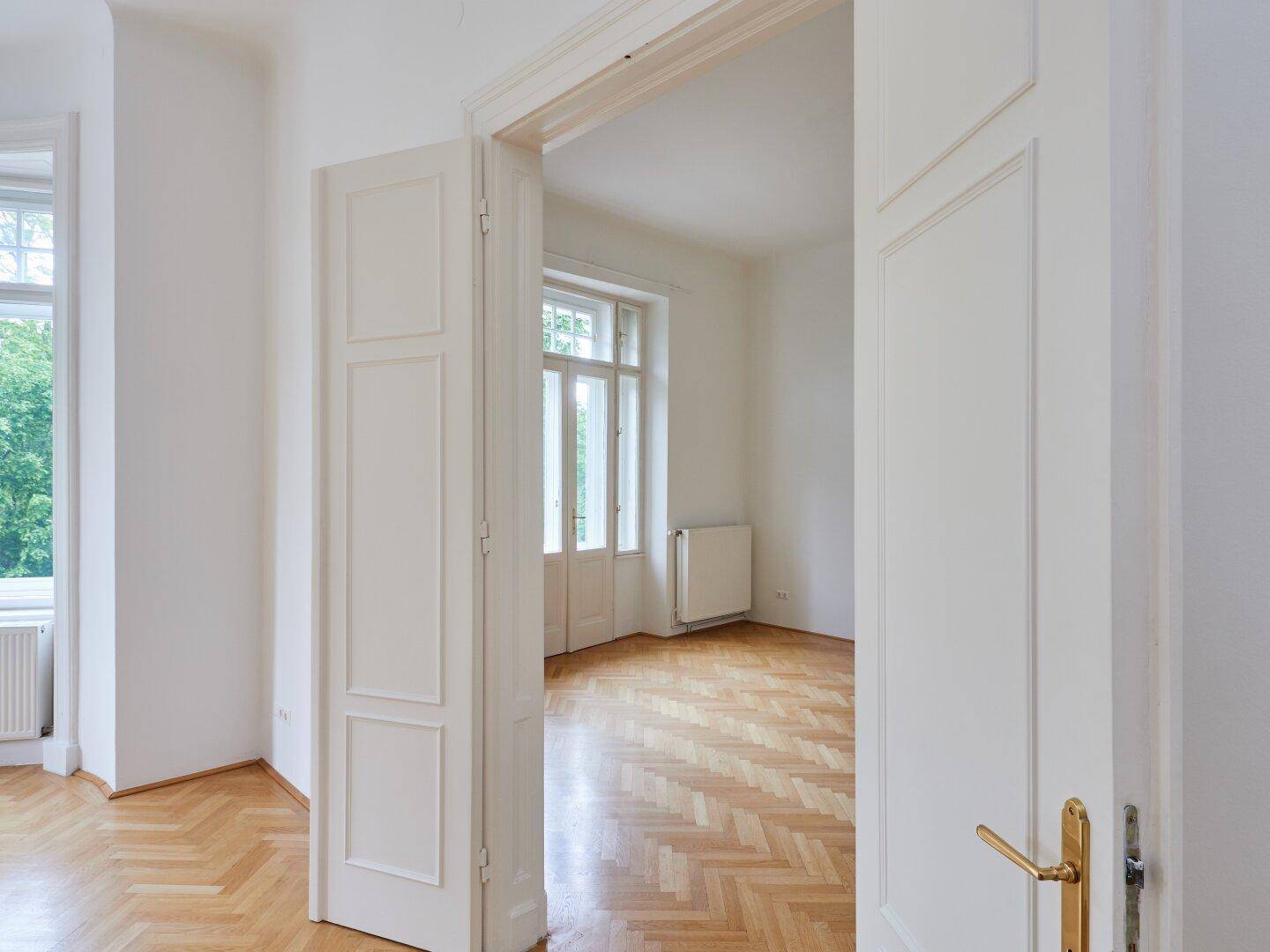 Offene Tür führt in ein weiteres helles Zimmer mit Fischgrätparkett und hohen Fenstern.