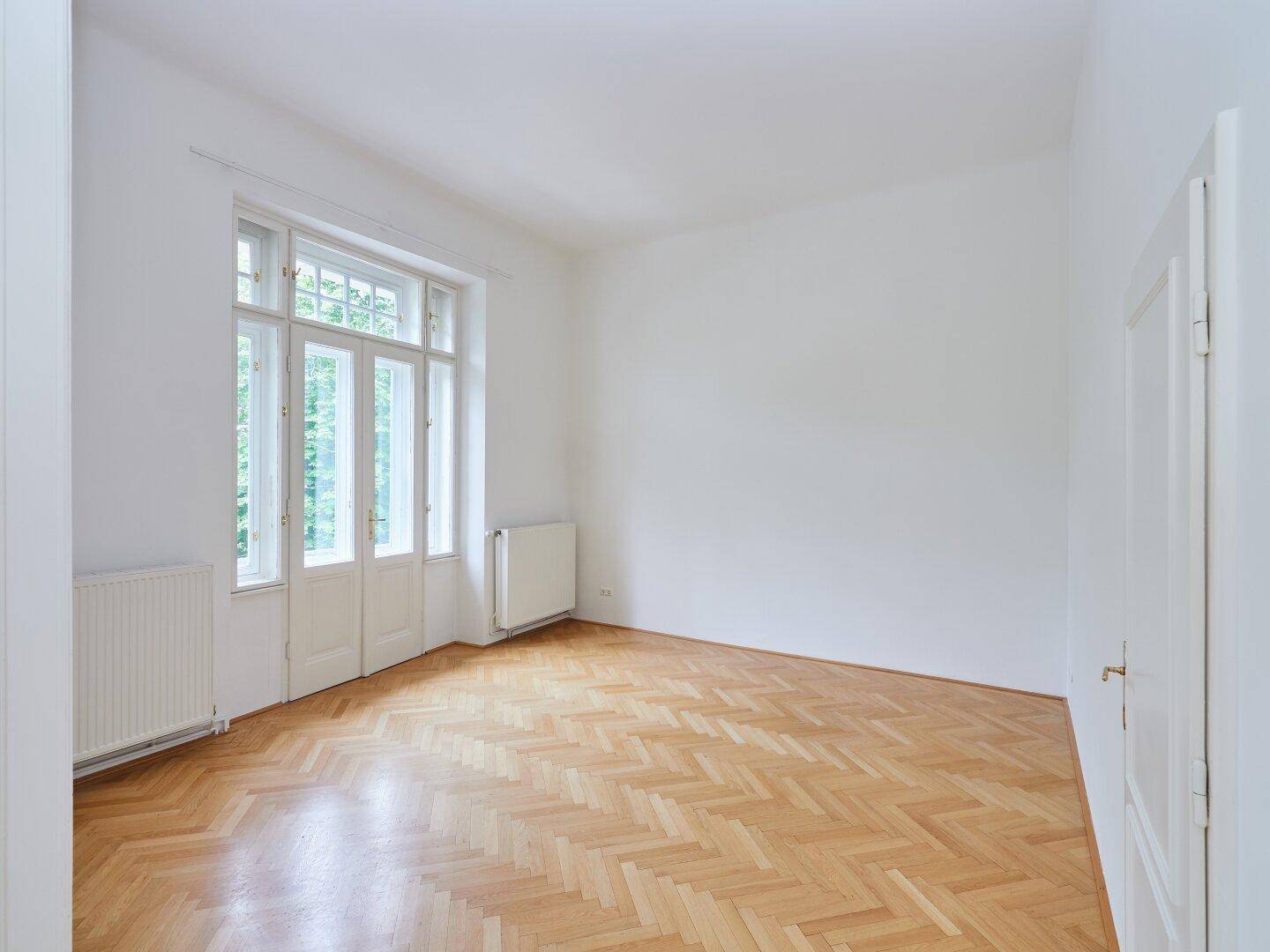 Helles Zimmer mit Fischgrätparkett und hohen Fenstern, die viel Tageslicht hereinlassen.