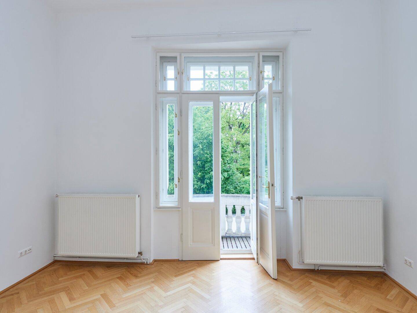 Helles Zimmer mit Fischgrätparkett und Zugang zu einem französischen Balkon mit Blick ins Grüne.