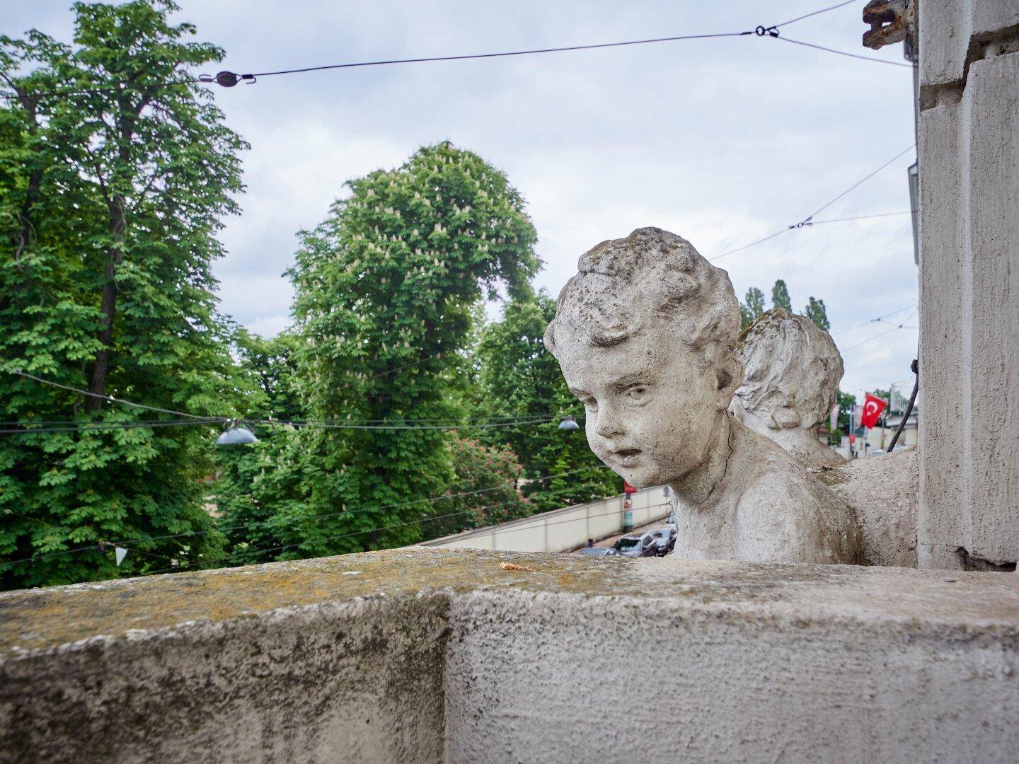 Detailansicht einer steinernen Balustrade mit Skulptur und Blick auf grüne Bäume.