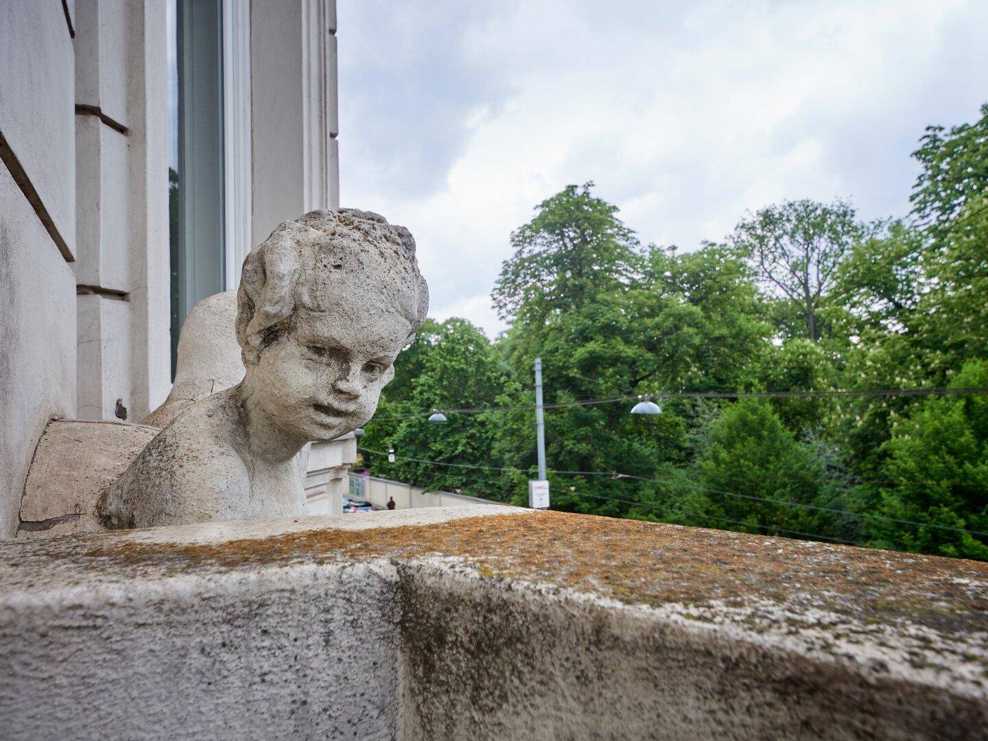 Steinerne Balustrade mit Skulptur und üppigem Grün im Hintergrund, unter bewölktem Himmel.