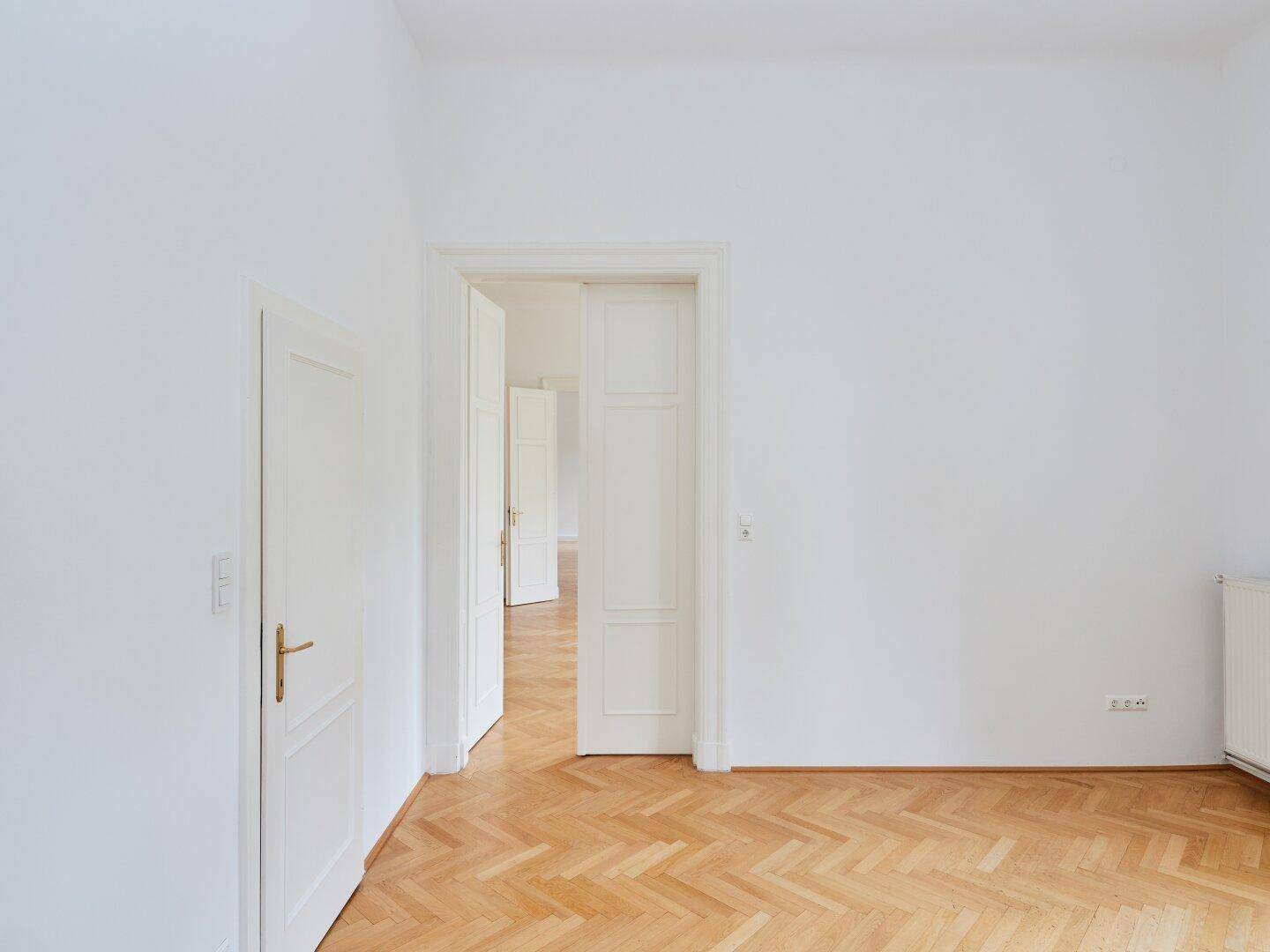 Leeres Zimmer mit Fischgrätparkett und weißen Türen, die zu weiteren Räumen führen.