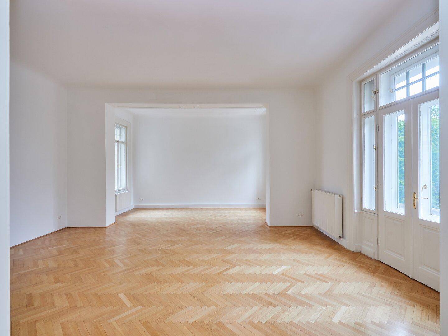 Geräumiges, helles Zimmer mit Fischgrätparkett und einem breiten Durchgang zu einem weiteren Bereich.