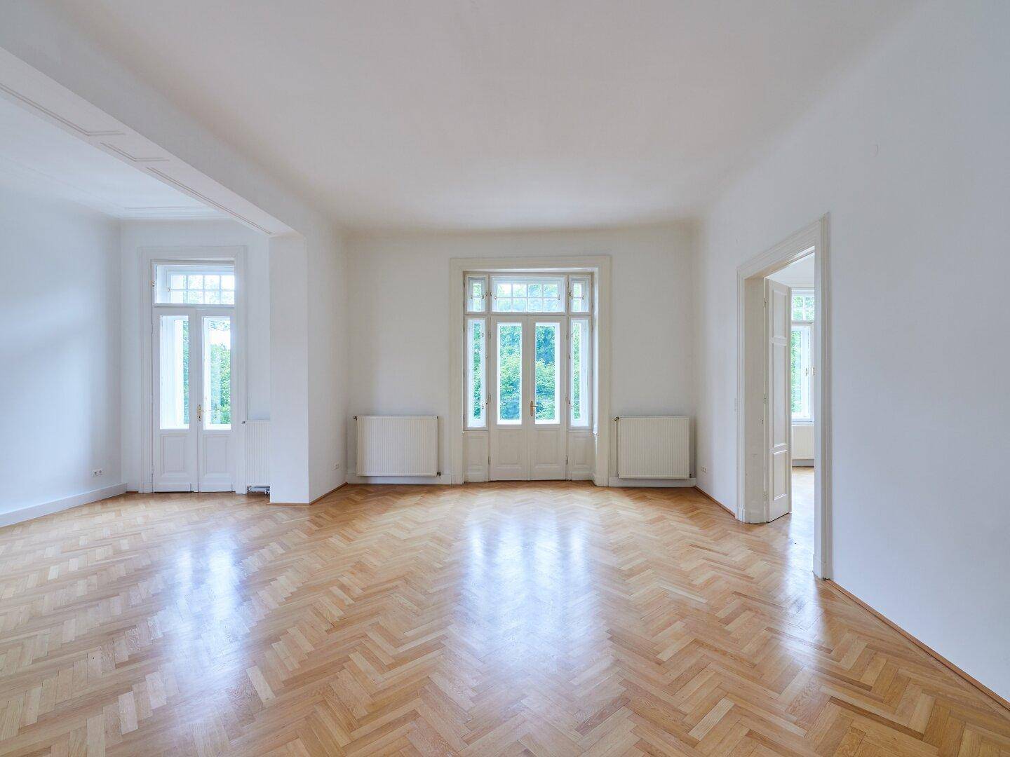 Großes, helles Zimmer mit Fischgrätparkett und mehreren Fenstern, die viel Licht spenden.