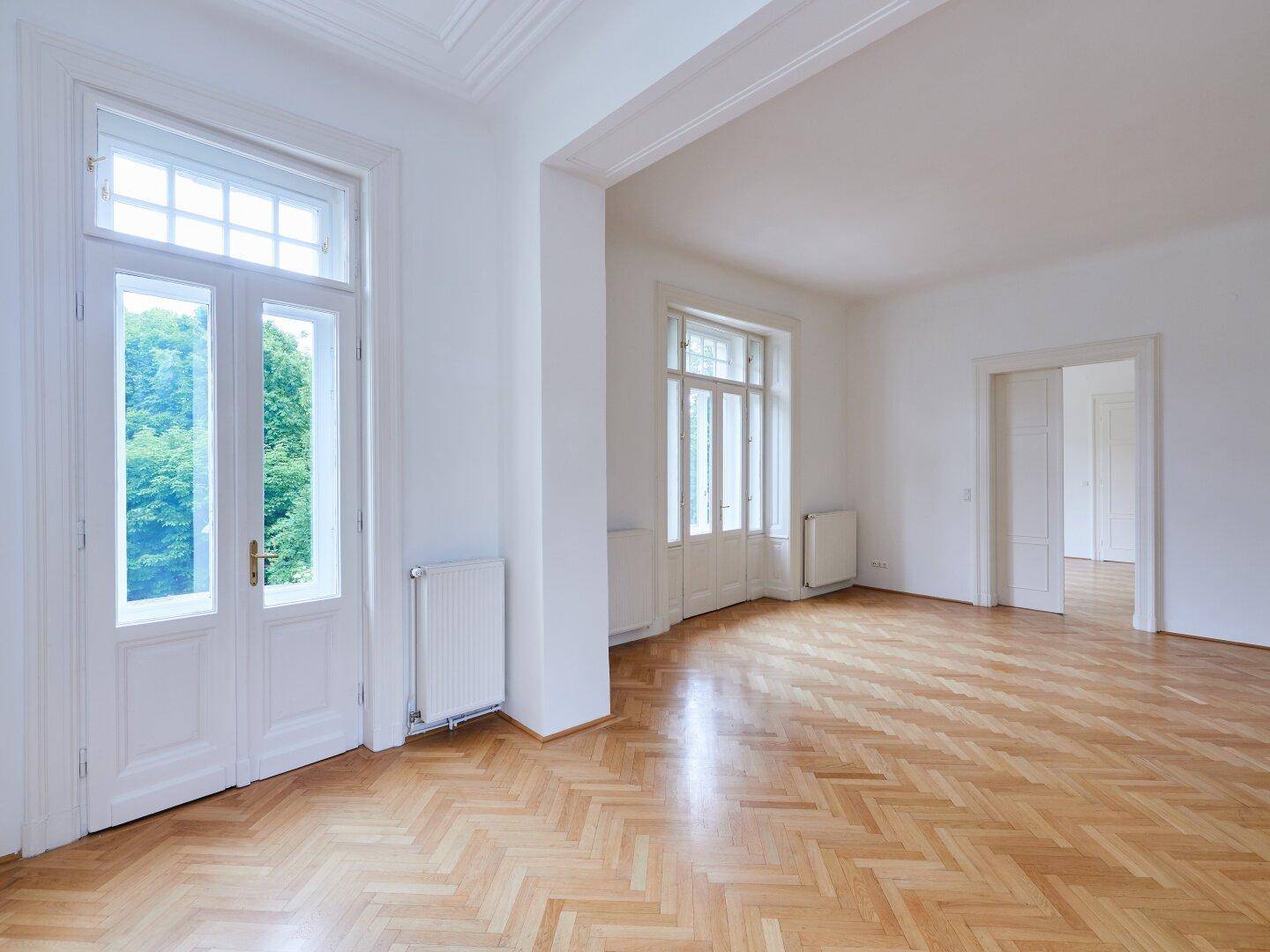 Helles Zimmer mit Fischgrätparkett und hohen Fenstern, die einen Blick ins Grüne ermöglichen.