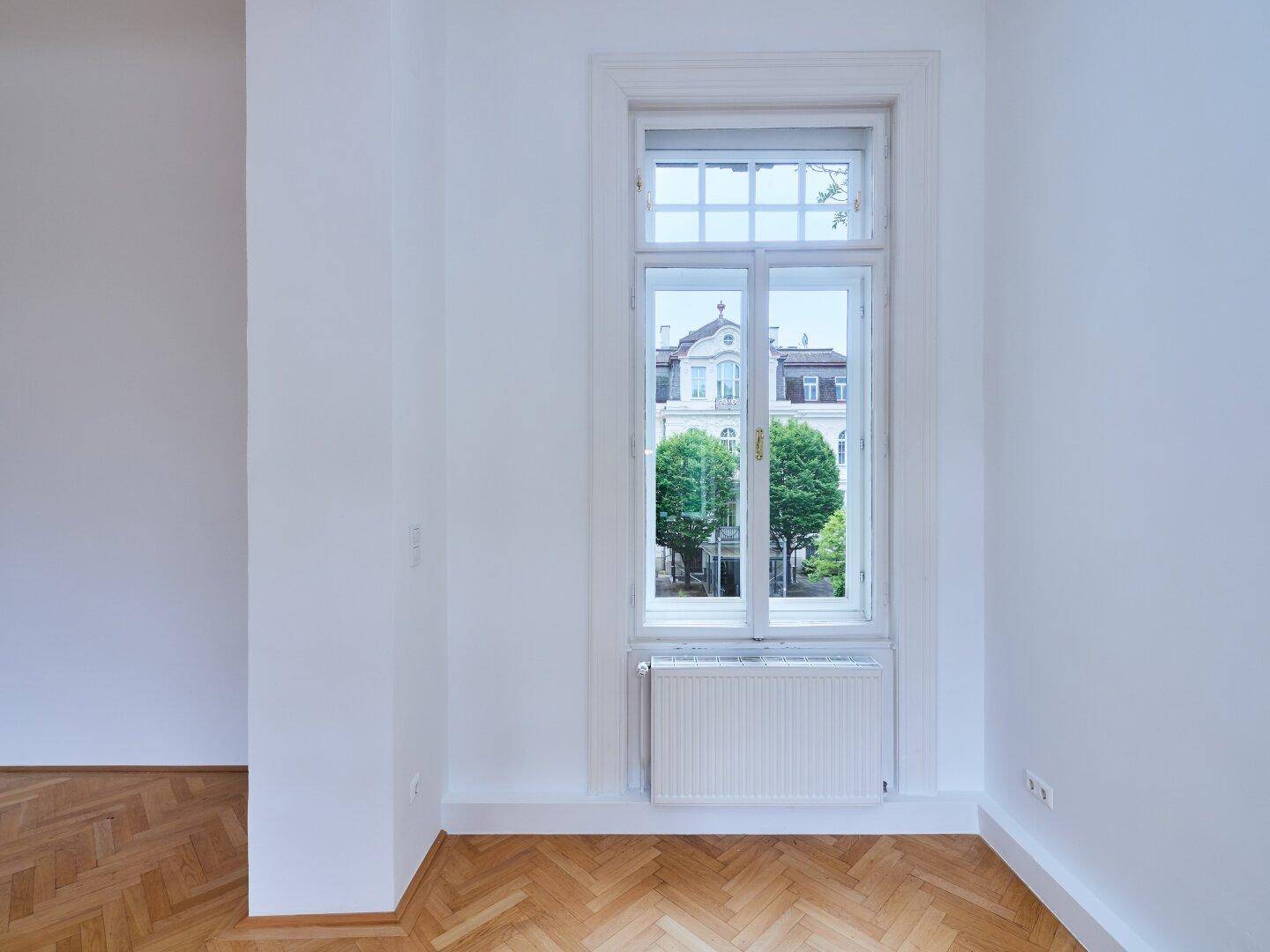 Helles Zimmer mit Fischgrätparkett und einem hohen Fenster mit Blick auf die Stadt.