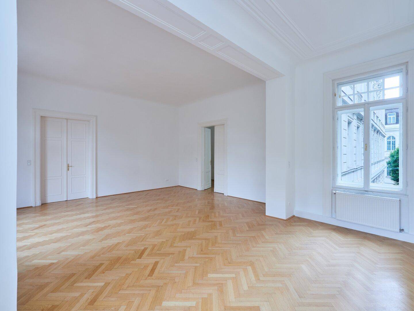 Geräumiges, helles Zimmer mit Fischgrätparkett und hohen Fenstern, die viel Tageslicht hereinlassen.