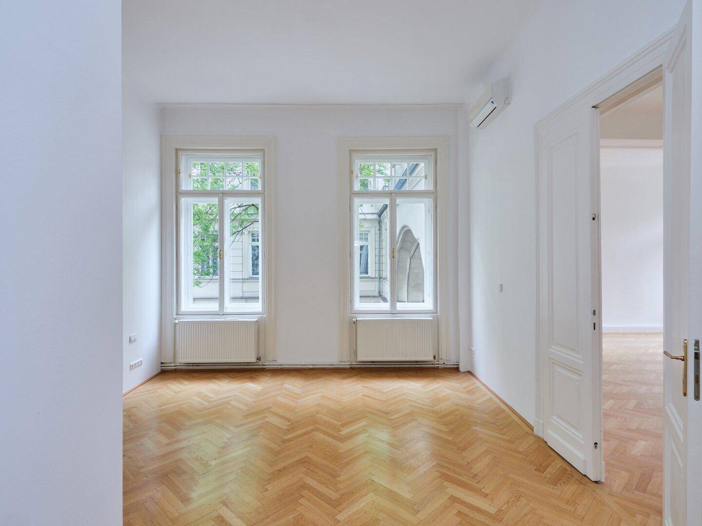 Helles Zimmer mit Fischgrätparkett, zwei hohen Fenstern und einer Klimaanlage an der Wand.