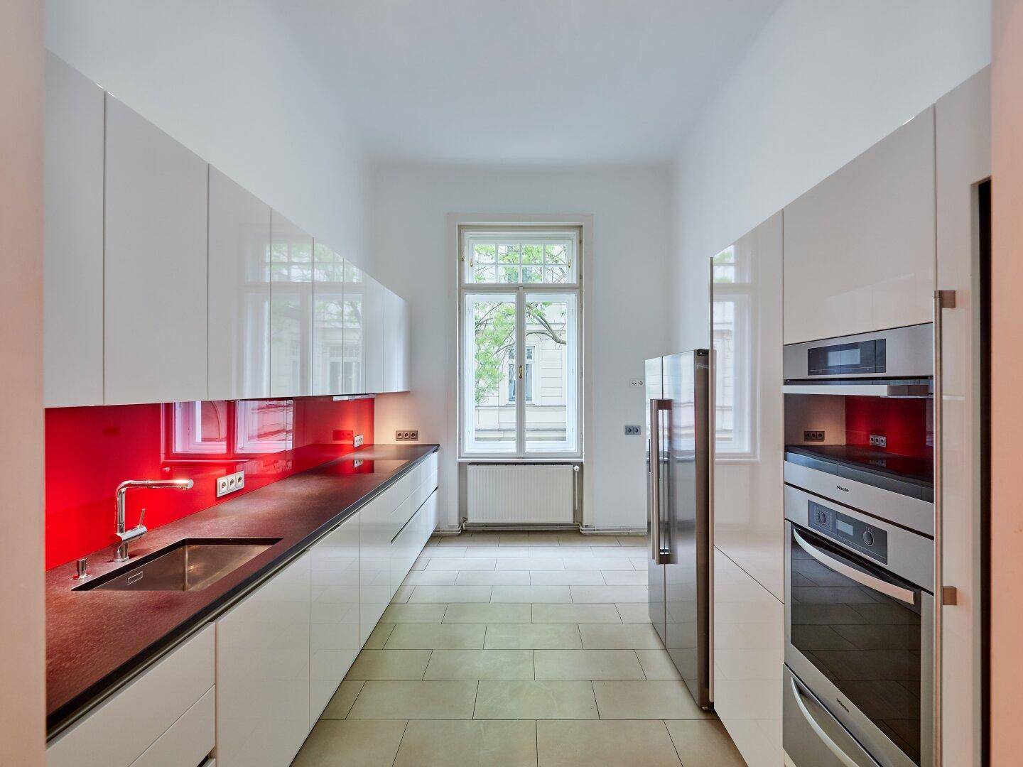 Lange, moderne Küche mit weißen Hochglanzschränken, roter Rückwand und Fenster.