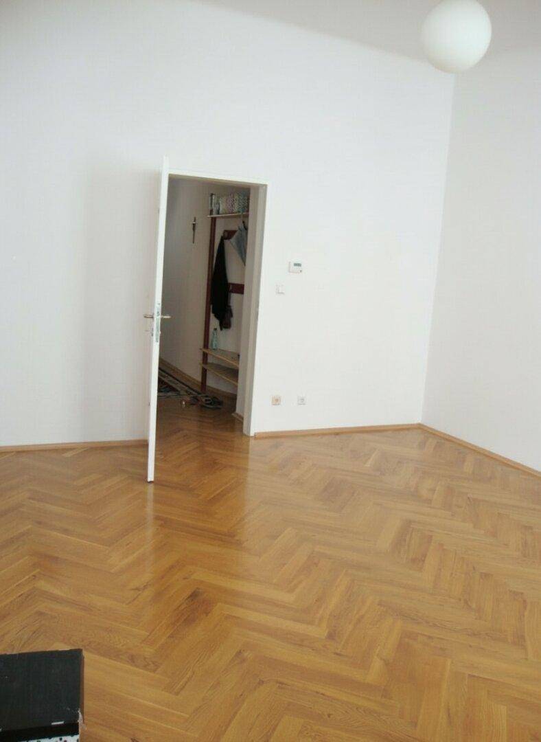 Leeres Zimmer mit glänzendem Fischgrätparkett und einer offenen Tür, die zu einem weiteren Raum führt.