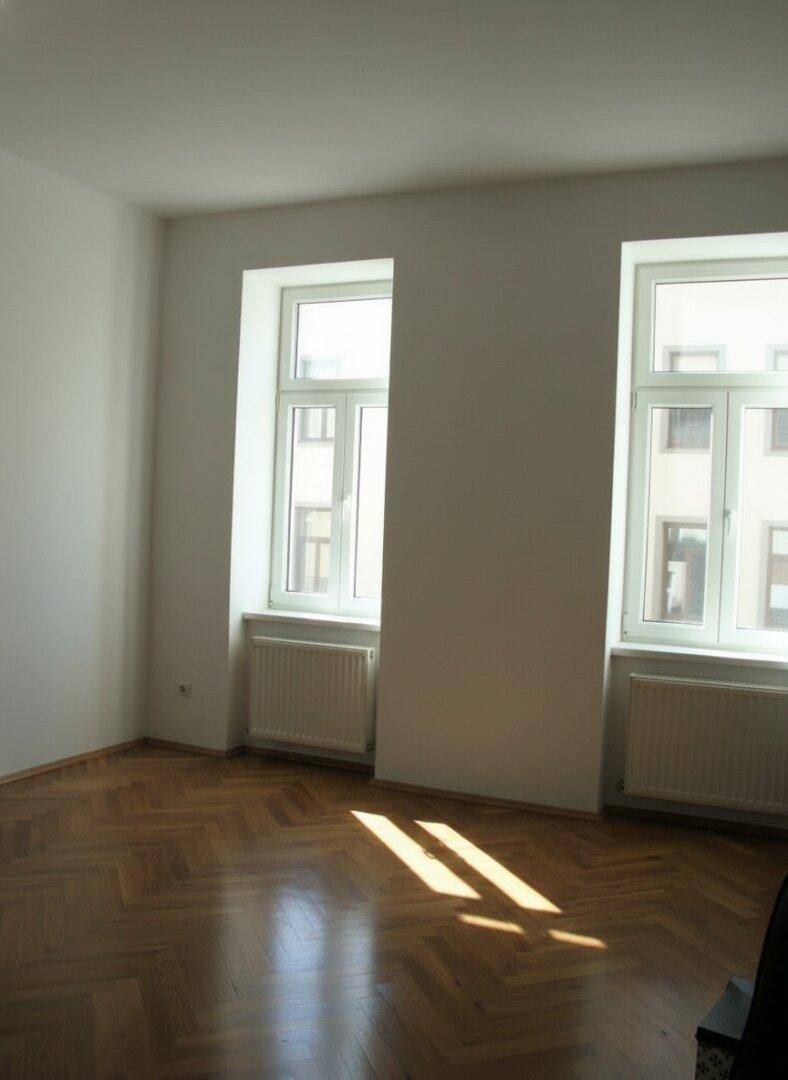 Sonniger Raum mit Fischgrätparkett und zwei Fenstern, durch die Sonnenstrahlen auf den Boden fallen.