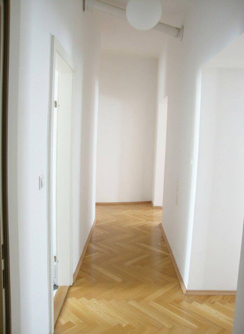 Langer, heller Flur mit Fischgrätparkettboden und weißen Wänden, der zu weiteren Räumen führt.