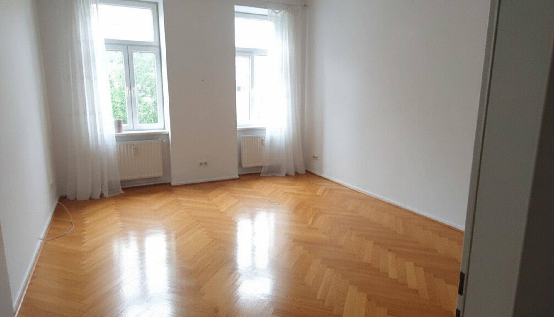 Helles Zimmer mit Fischgrätparkett, weißen Vorhängen an den Fenstern und viel Tageslicht.