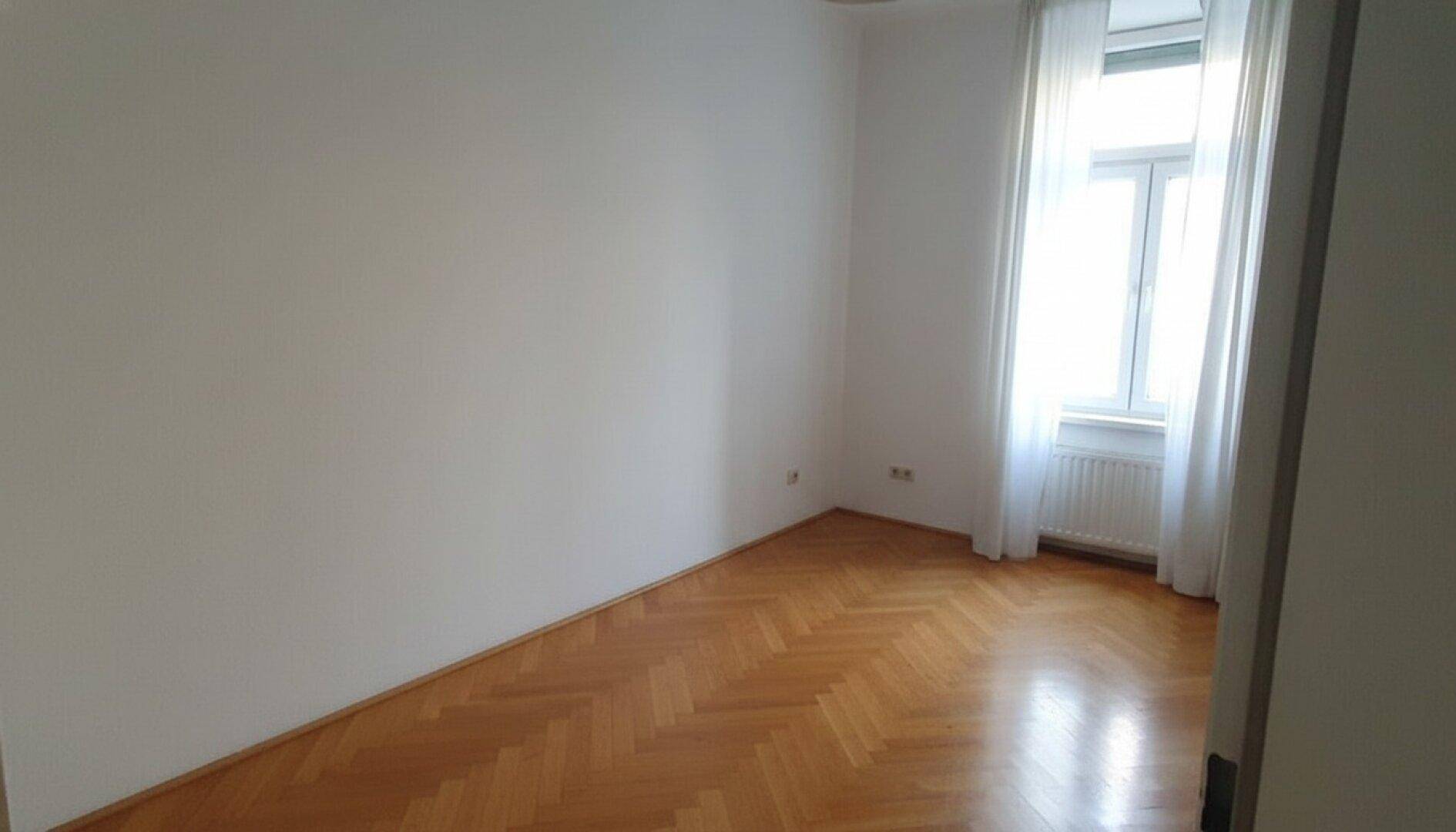 Leeres Zimmer mit glänzendem Fischgrätparkett und einem Fenster mit weißen Vorhängen.