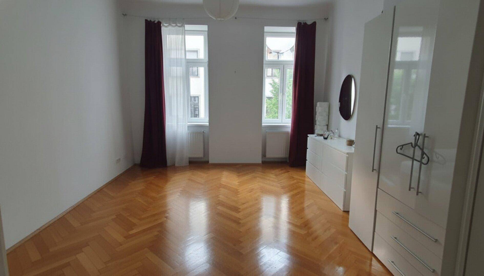 Schlafzimmer mit Fischgrätparkett, weißen Möbeln und roten Vorhängen an den beiden Fenstern.