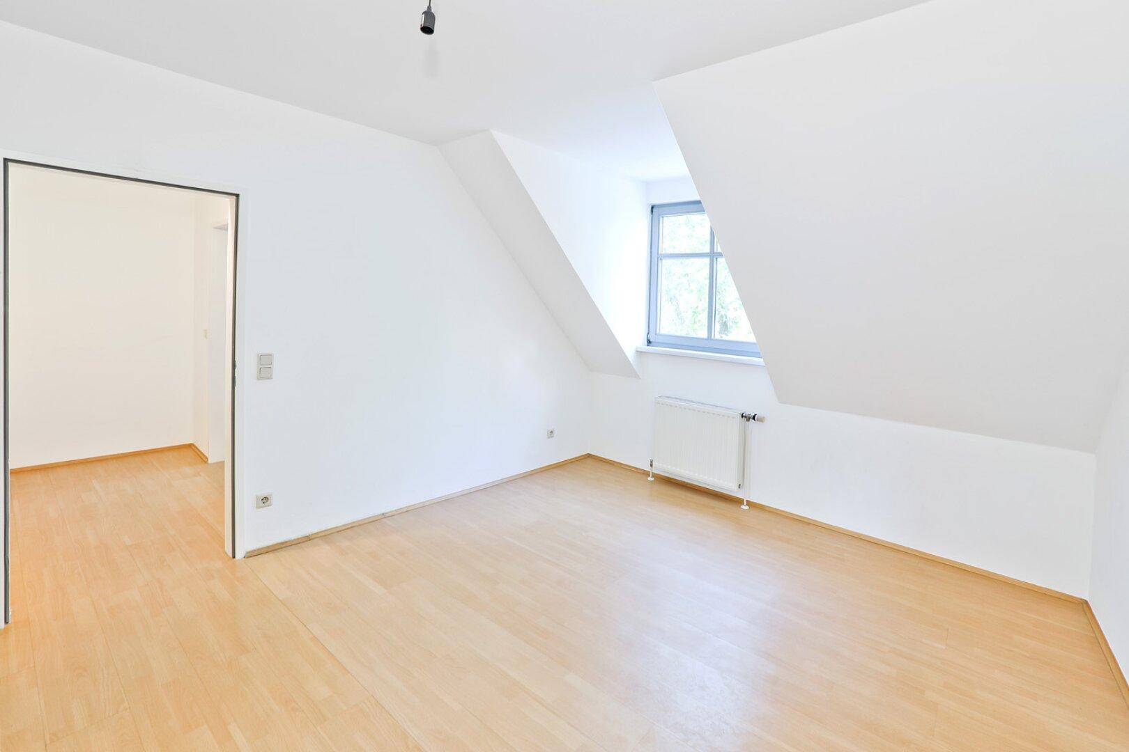 Helles Zimmer im Dachgeschoss mit Schrägen, Parkettboden und Fenster.