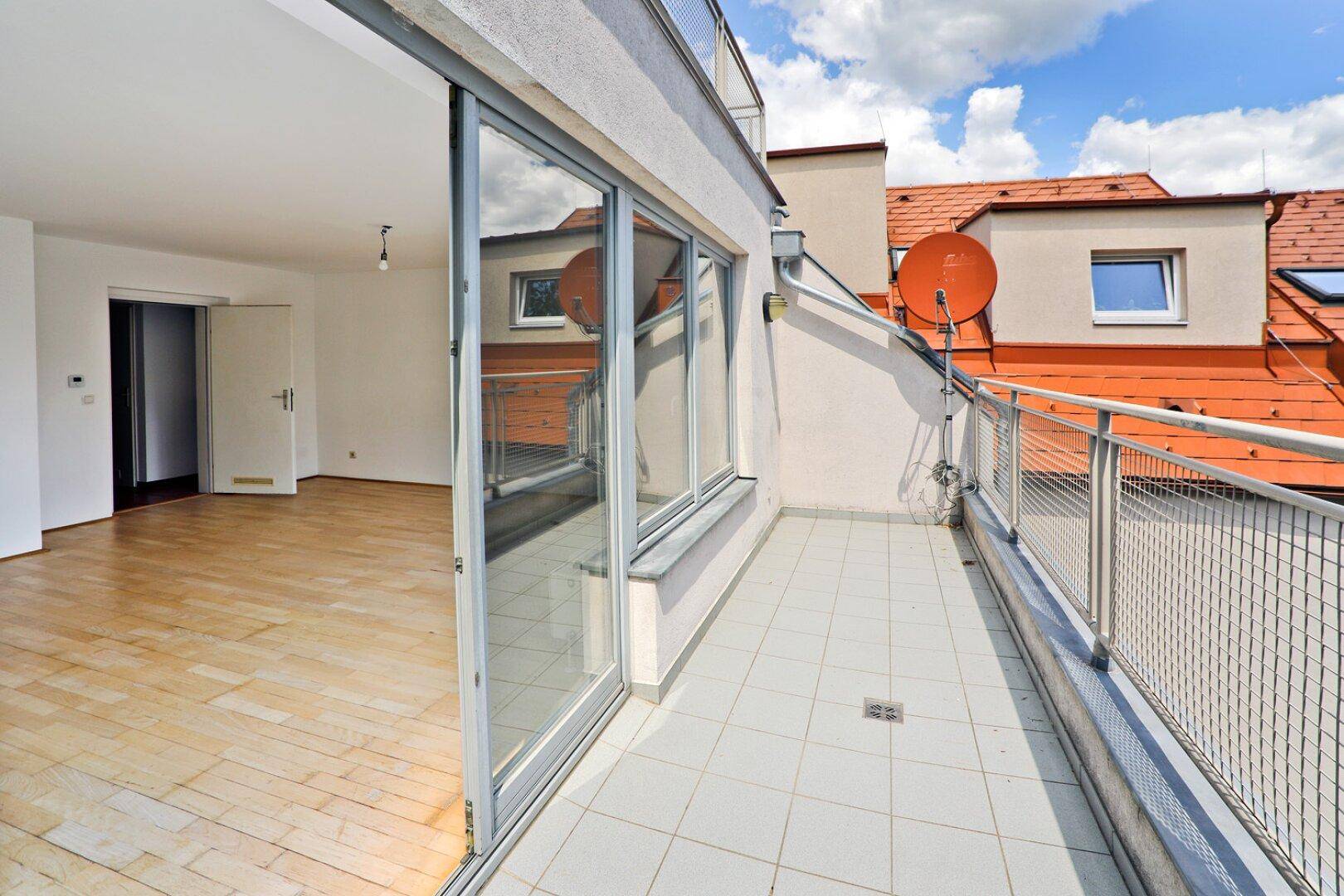 Geräumiger Balkon mit Fliesenboden und Blick auf die Dächer und Bäume.