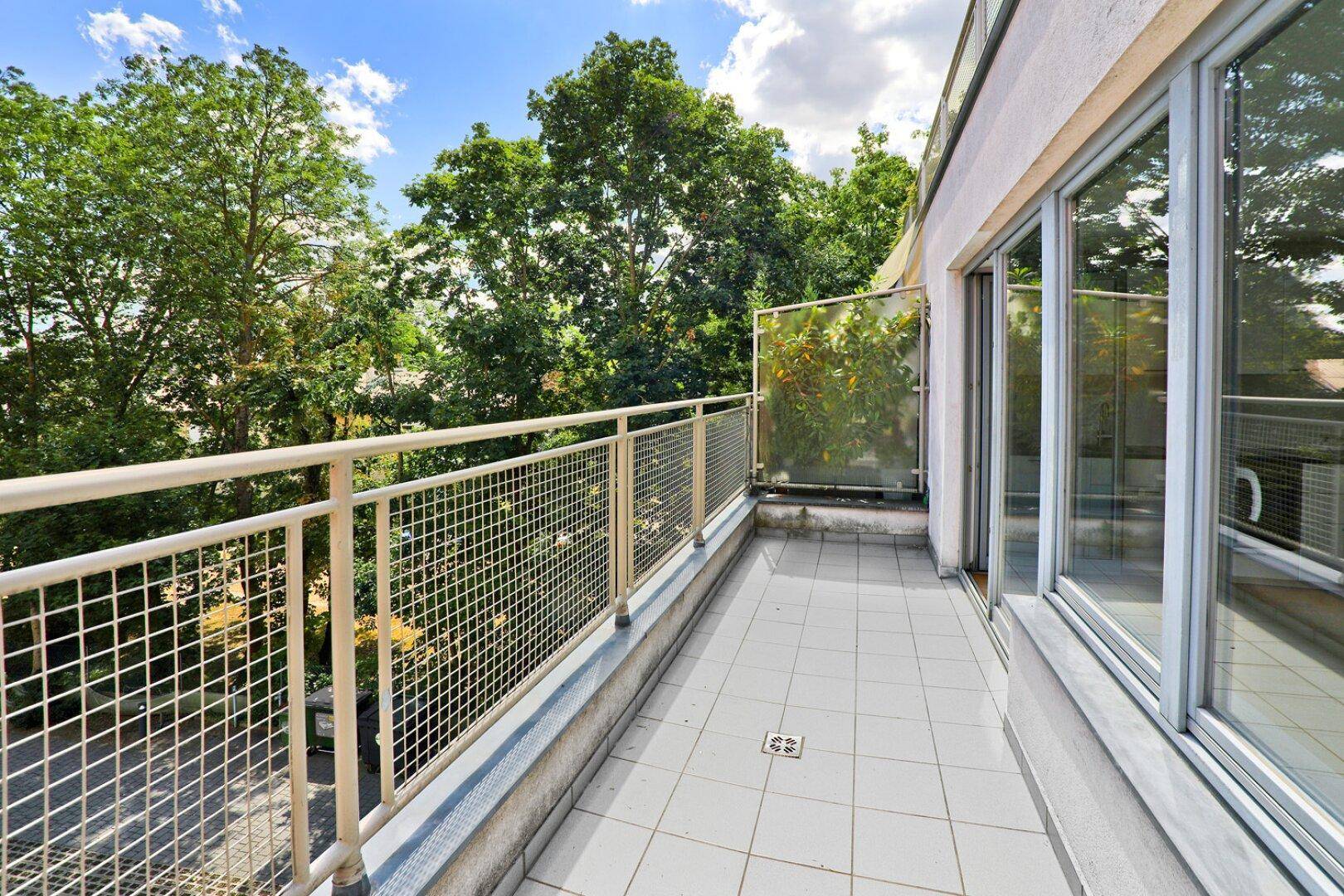 Heller Balkon mit Fliesenboden und Metallgeländer, umgeben von Bäumen.