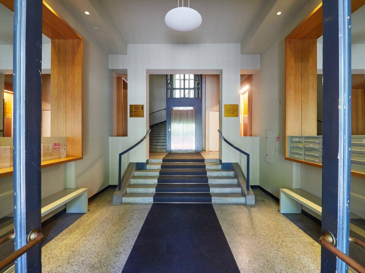 Heller Eingangsbereich mit Treppe, Aufzug und Terrazzoboden in einem eleganten Gebäude.