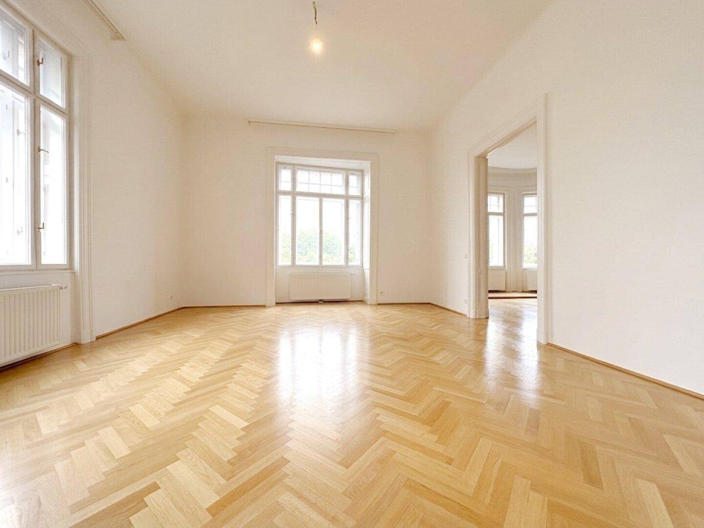 Helles Zimmer mit klassischem Fischgrätparkett und hohen Fenstern, das eine offene und einladende Atmosphäre bietet.