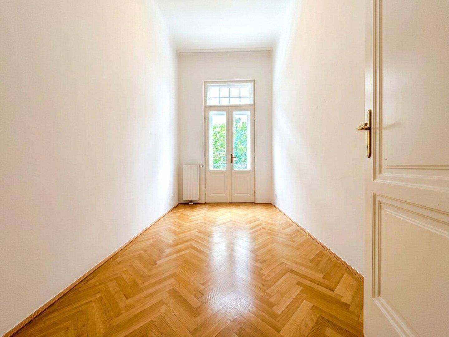 Schmaler, heller Raum mit Fischgrätparkett und einer Doppeltür, die ins Freie führt.