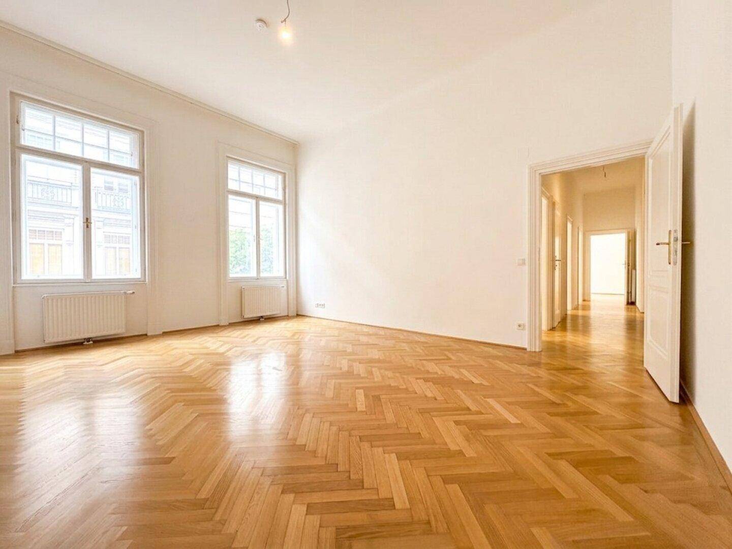 Großes, helles Zimmer mit Fischgrätparkett und hohen Fenstern, das einen Blick in den Flur bietet.