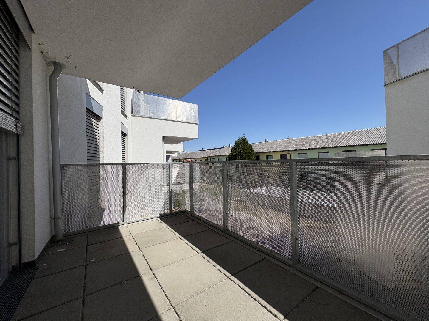 Sonniger Balkon mit Fliesenboden und moderner Metallbrüstung, umgeben von anderen Gebäuden.