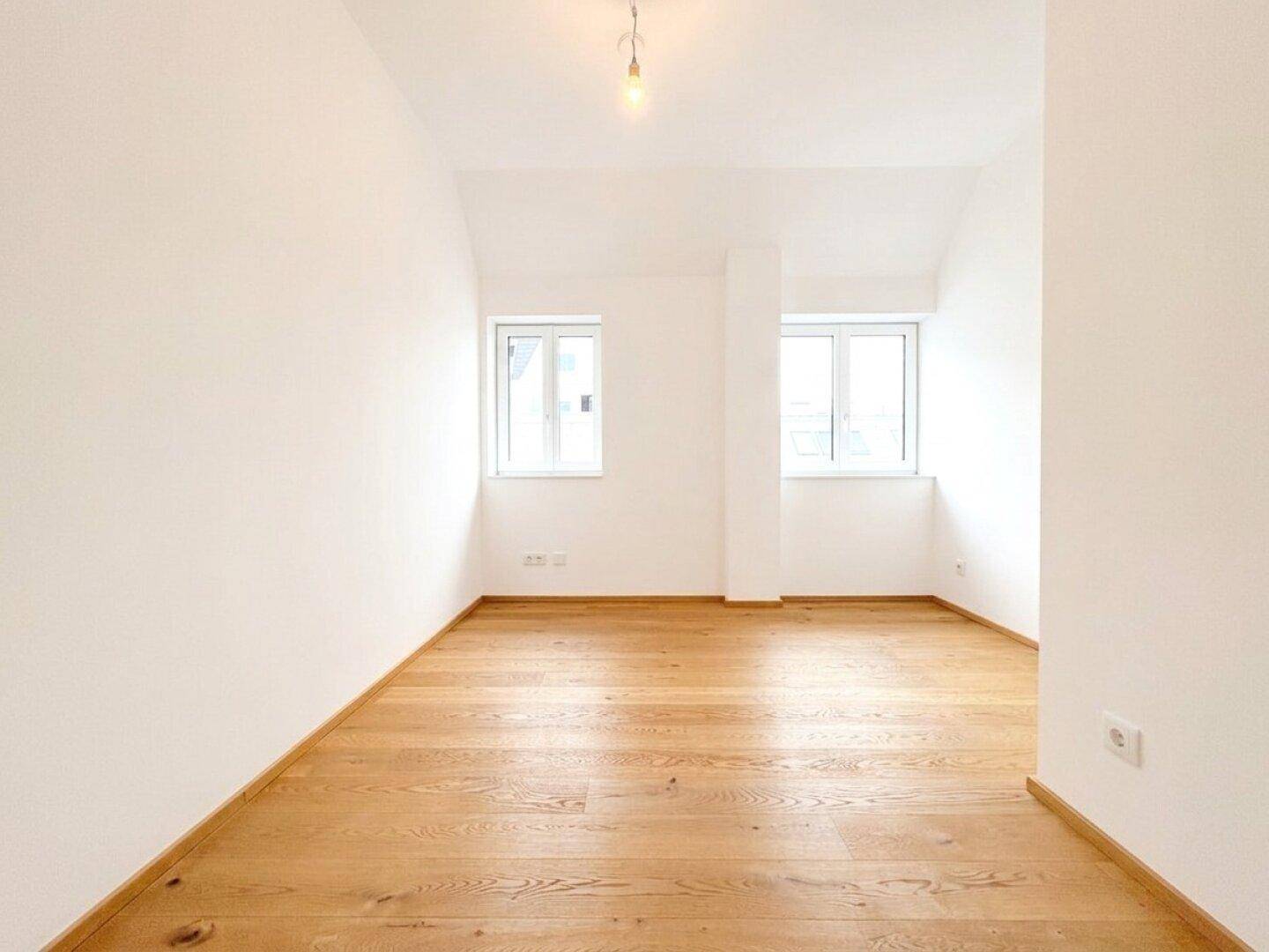 Leeres Schlafzimmer mit hellem Holzboden, weißen Wänden und zwei Fenstern für viel Licht.