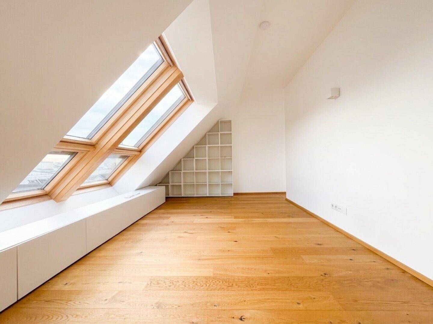 Lichtdurchfluteter Dachgeschossraum mit Holzboden, schrägen Wänden und praktischem Regal.