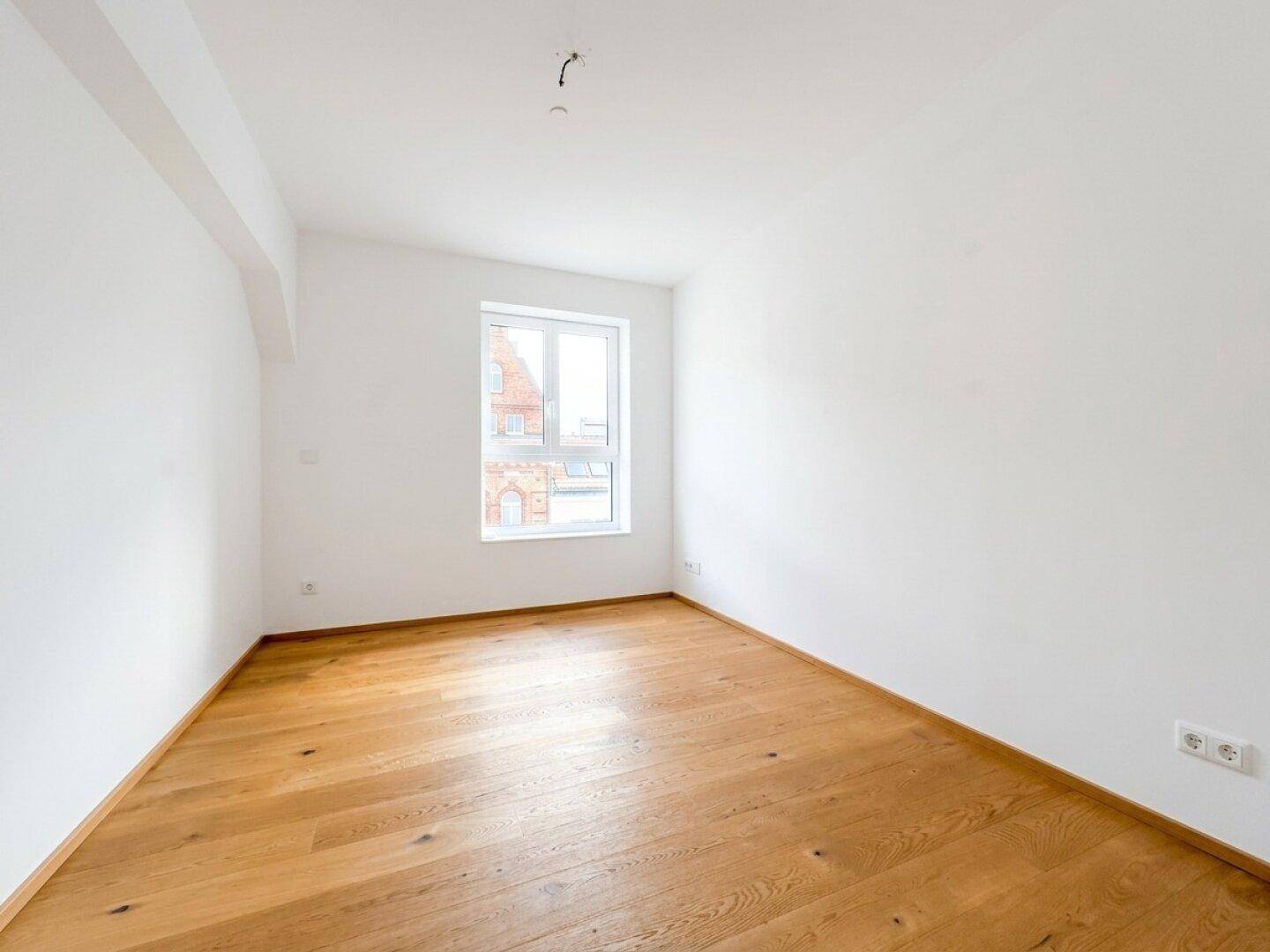 Leeres Schlafzimmer mit Holzboden, weißen Wänden und einem großen Fenster.